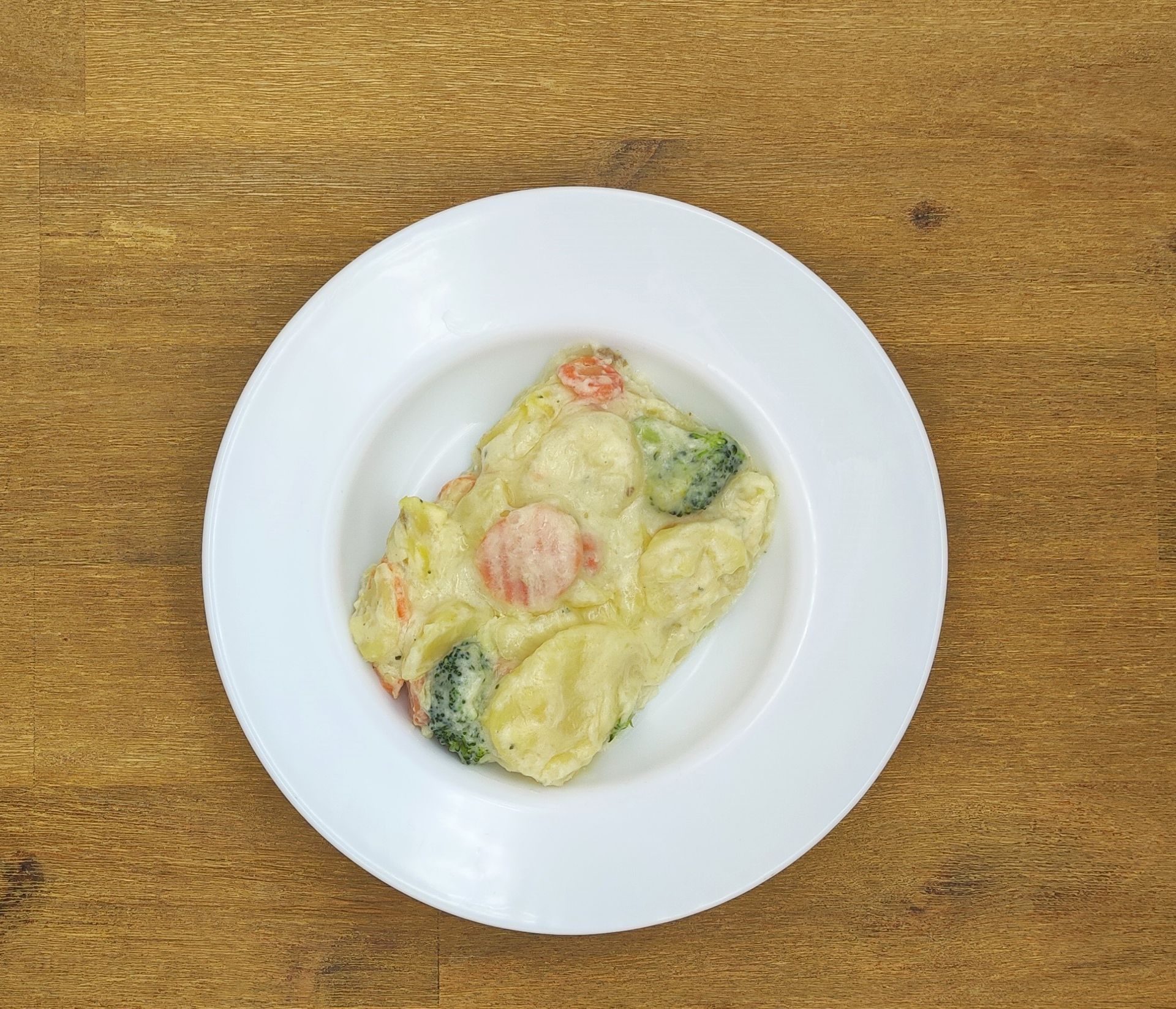 Cremiger Auflauf mit Brokkoli und Karotten in einer weißen Schüssel auf einem Holztisch.