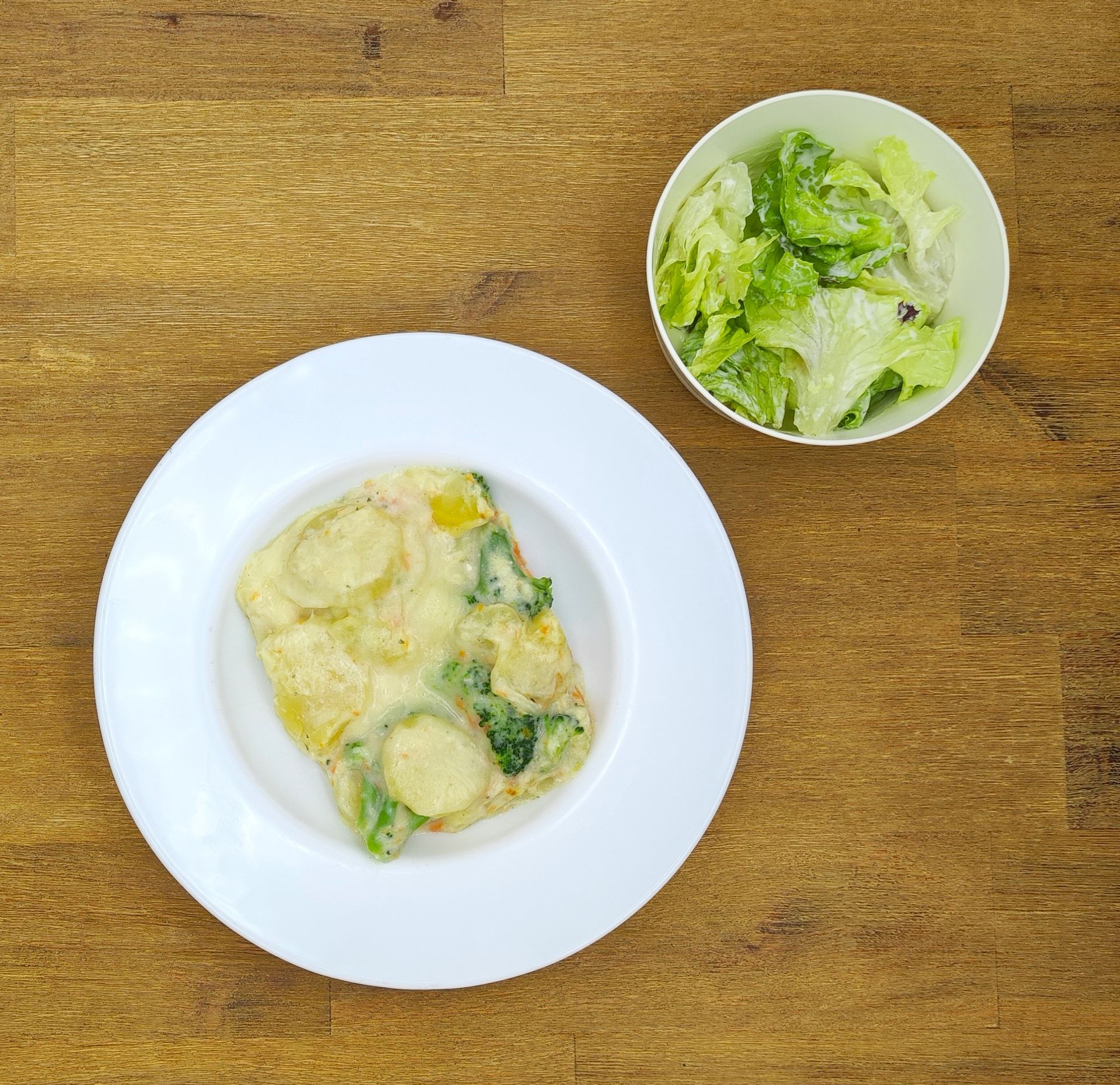 Omelett mit Kartoffeln und Brokkoli auf einem weißen Teller, dazu ein Beilagensalat in einer weißen Schüssel. Holztisch.