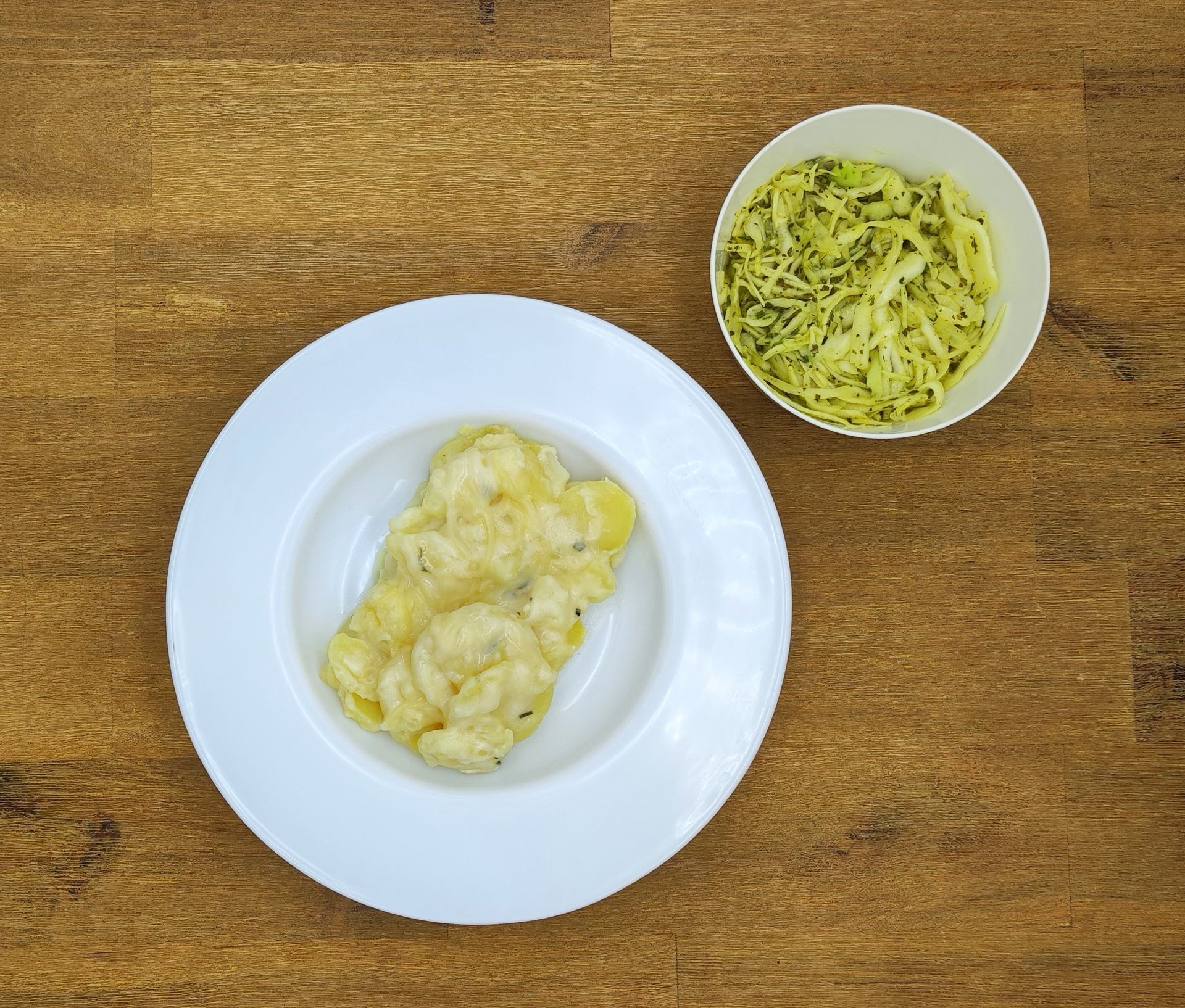 Ein weißer Teller mit cremigen Kartoffeln und eine Schüssel mit grünem Krautsalat auf einem Holztisch.