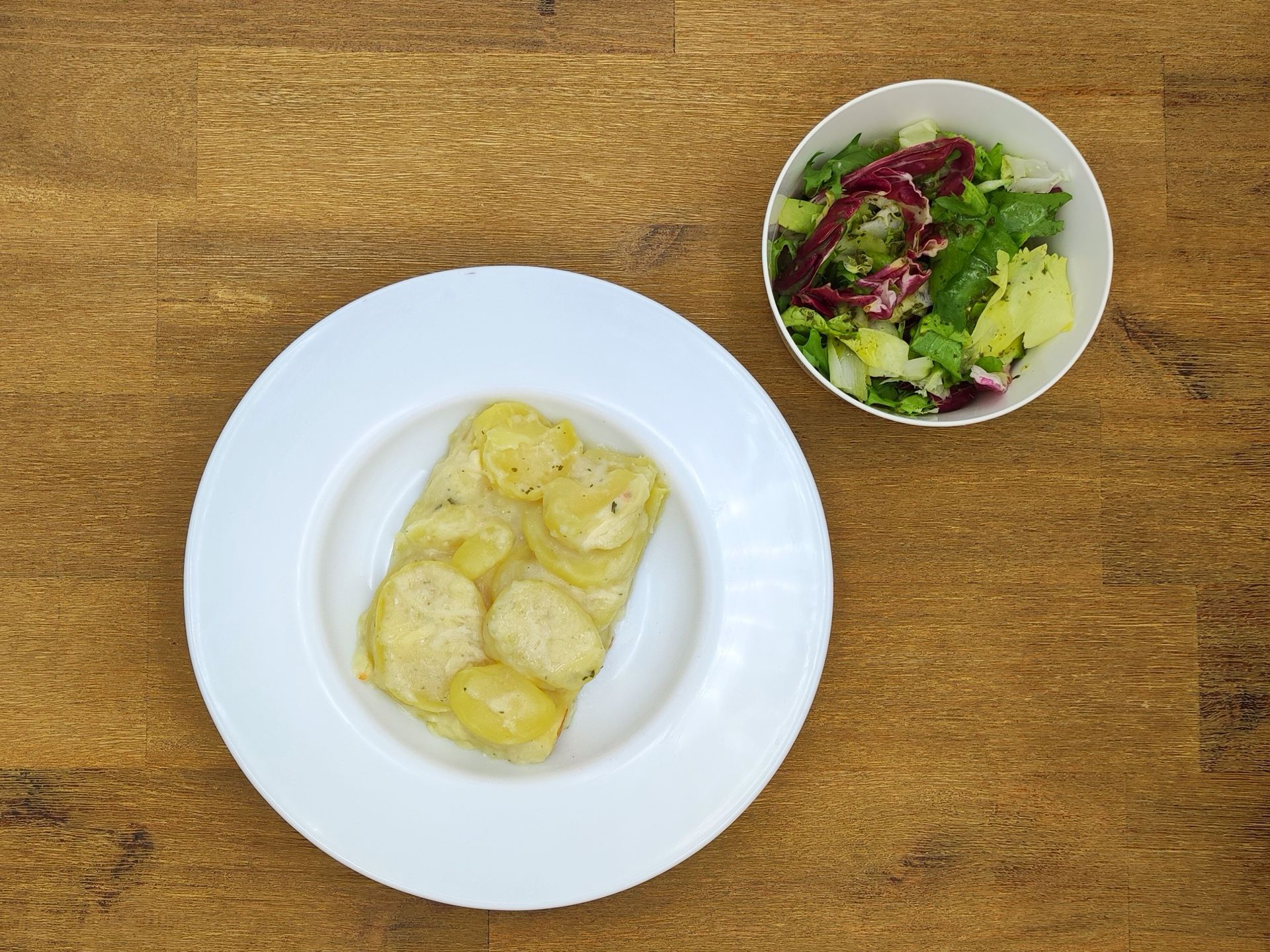 Kartoffelgratin auf einem weißen Teller mit einem Beilagensalat in einer weißen Schüssel, auf einer Holzoberfläche.