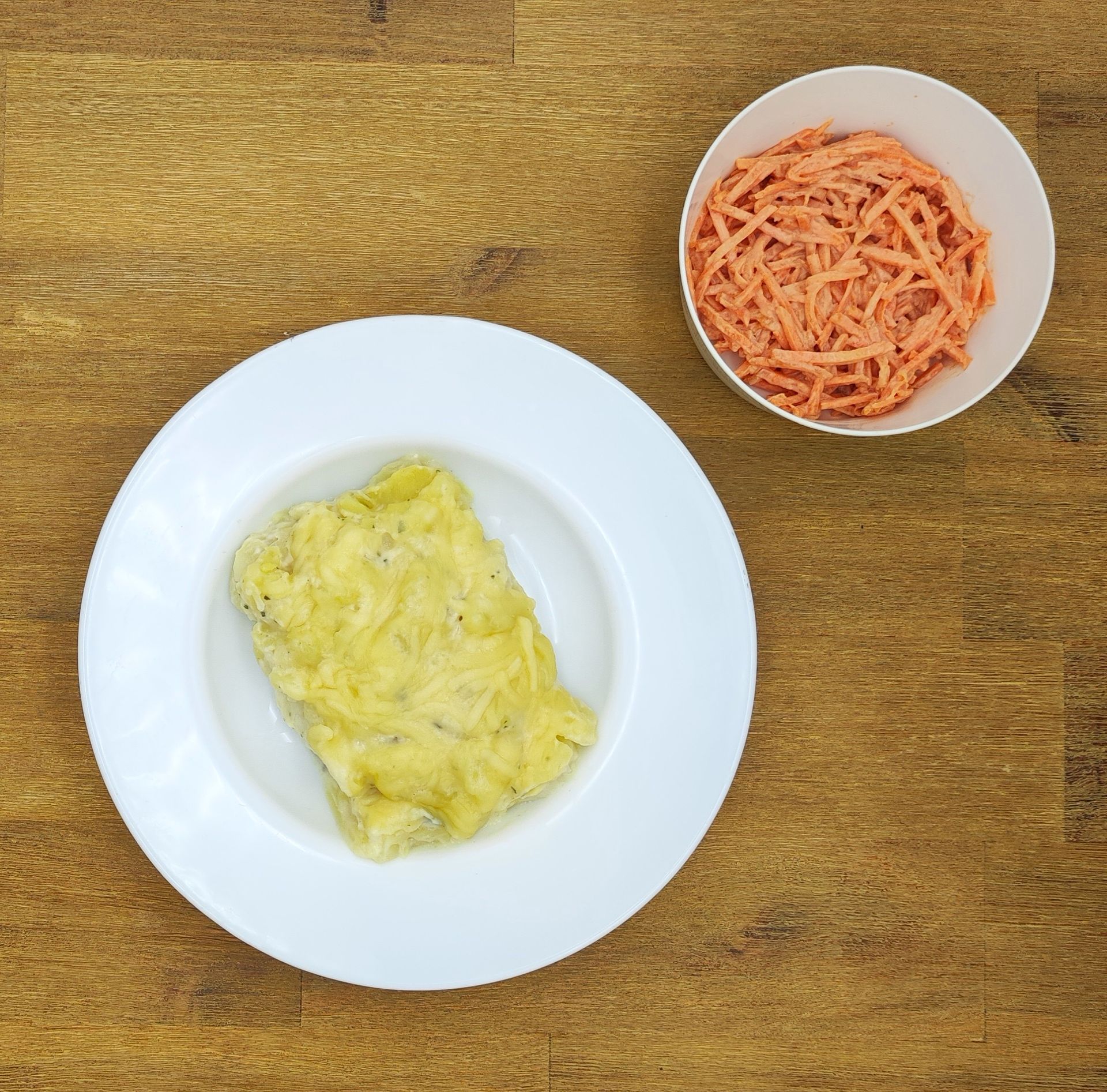 Weißer Teller mit cremiger Vorspeise neben einer Schüssel mit geraspelten Karotten auf einem Holztisch.