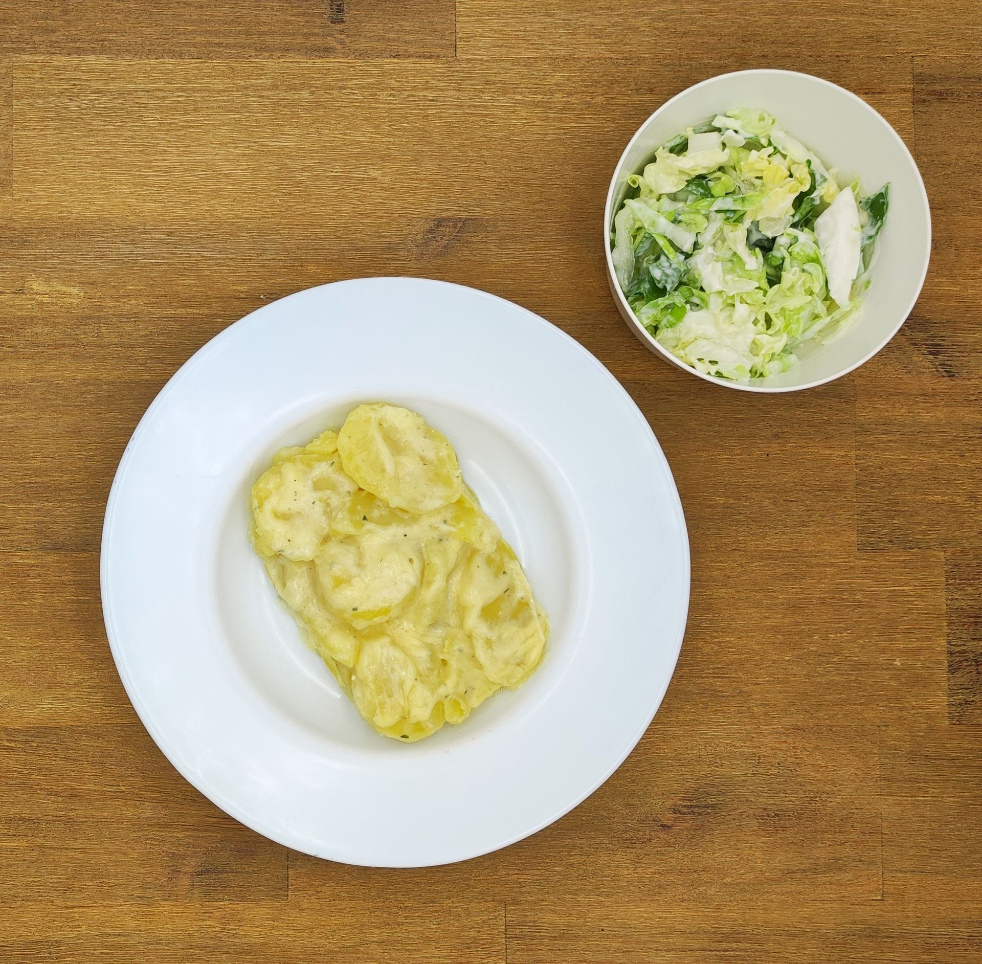 Kartoffelgratin auf einem weißen Teller, eine kleine Schüssel Salat auf einer Holzoberfläche.