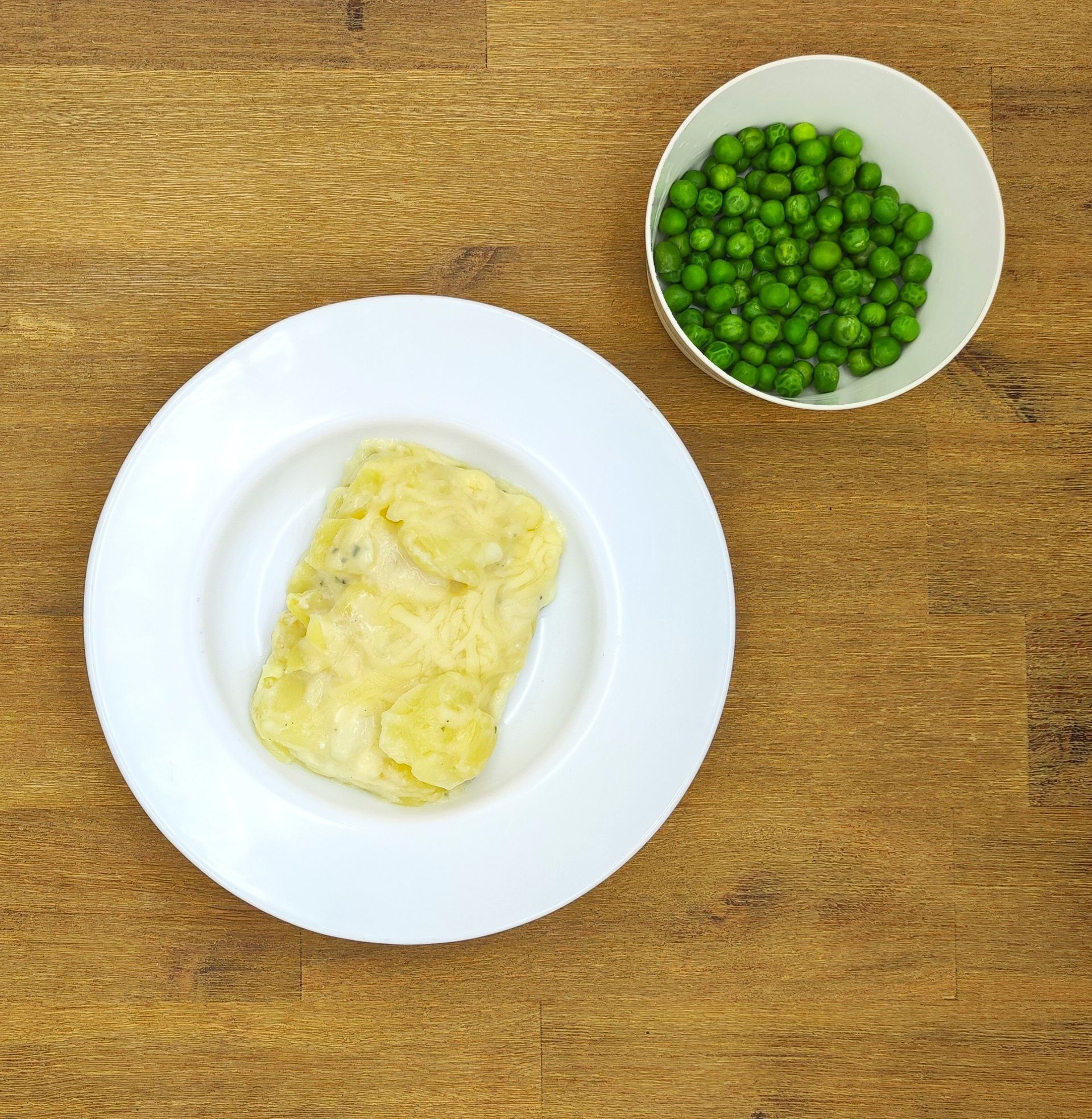 Weißer Teller mit Kartoffelgericht und Schüssel Erbsen auf einem Holztisch.