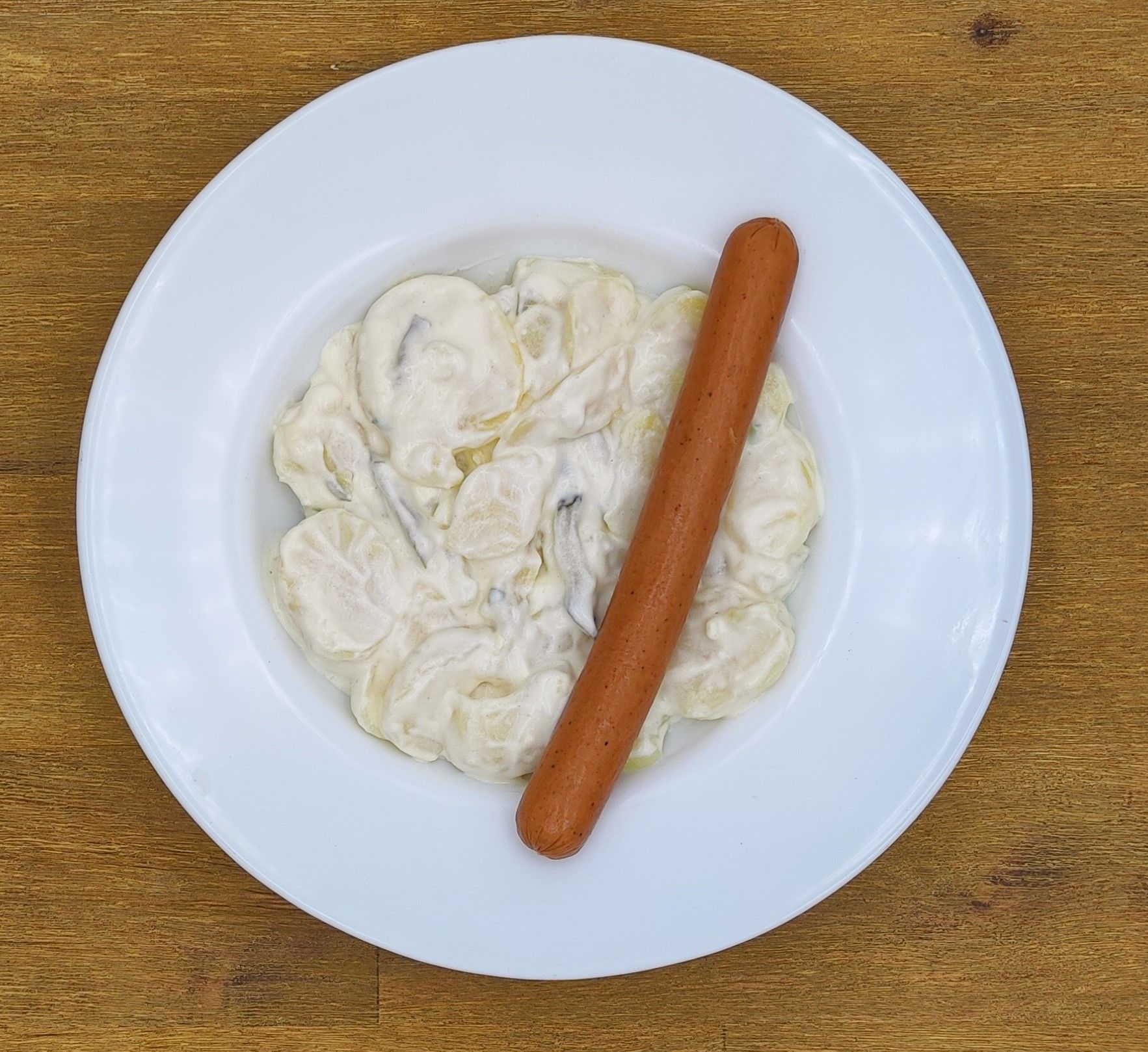 Weißer Teller mit Kartoffelsalat und einem Hotdog auf einem Holztisch.