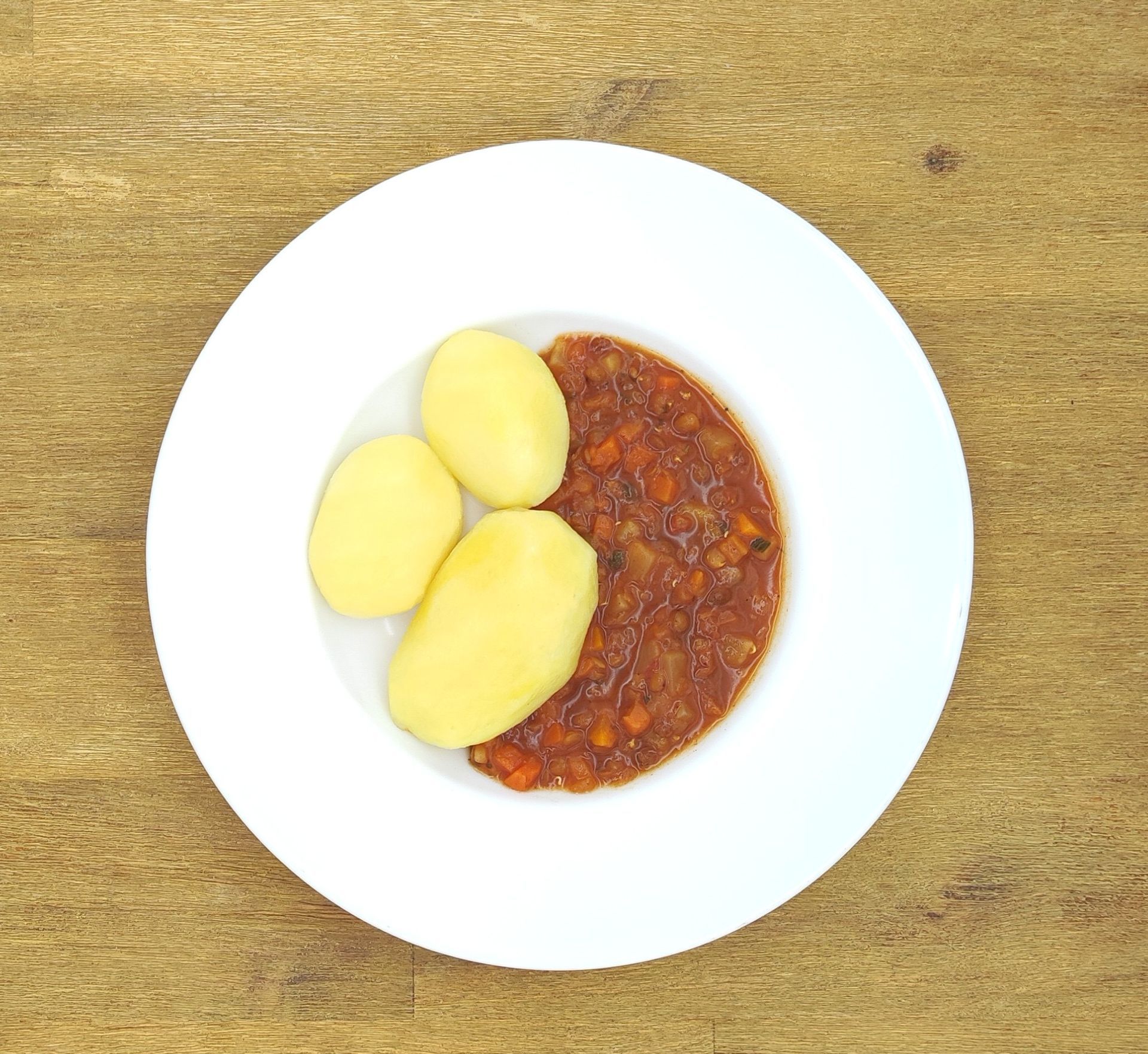Weißer Teller mit drei gekochten Kartoffeln und einem rotbraunen Eintopf auf einem Holztisch.