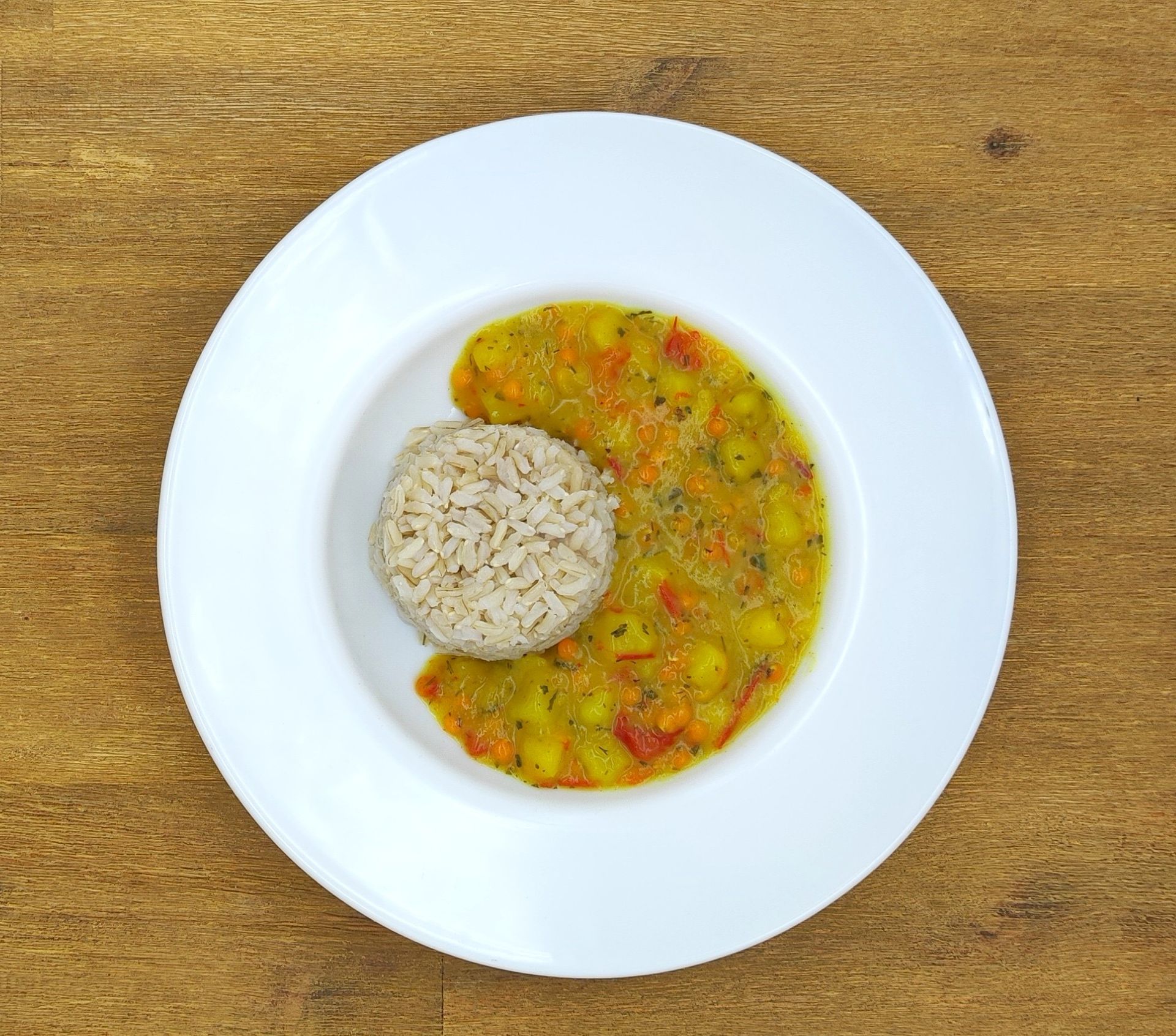 Weißer Teller mit gelbem Eintopf und braunem Reis auf einem Holztisch.