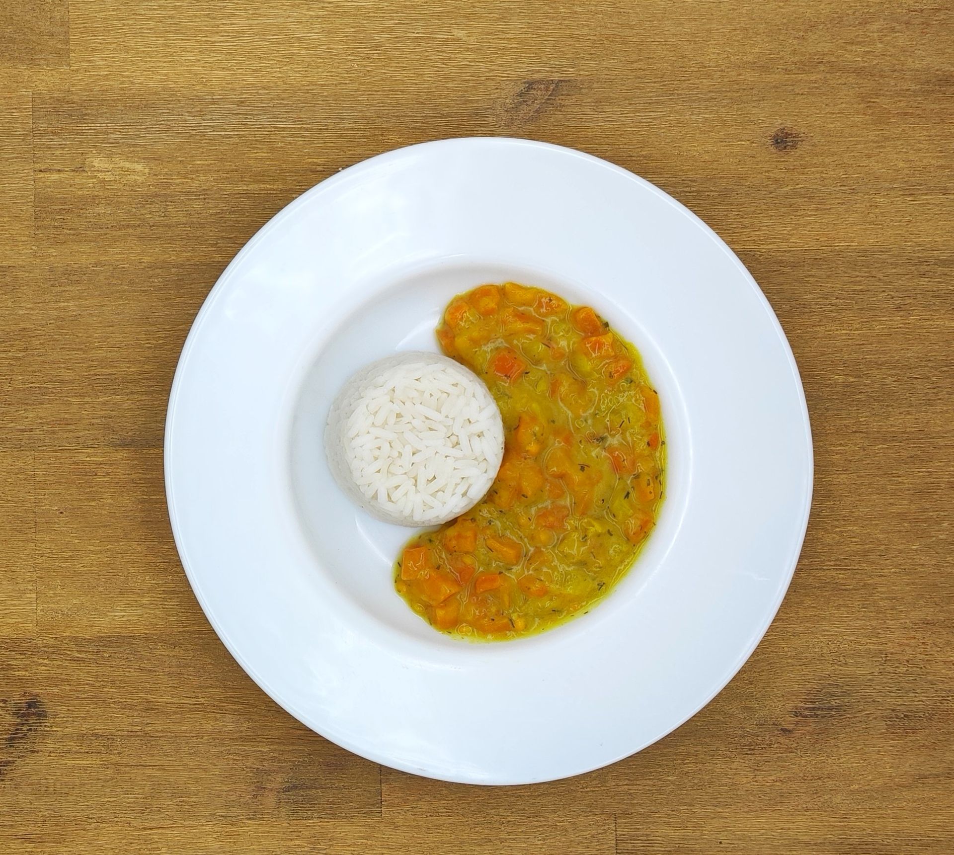 Weißer Teller mit einer Kugel Reis und Orangencurry auf einem Holztisch.