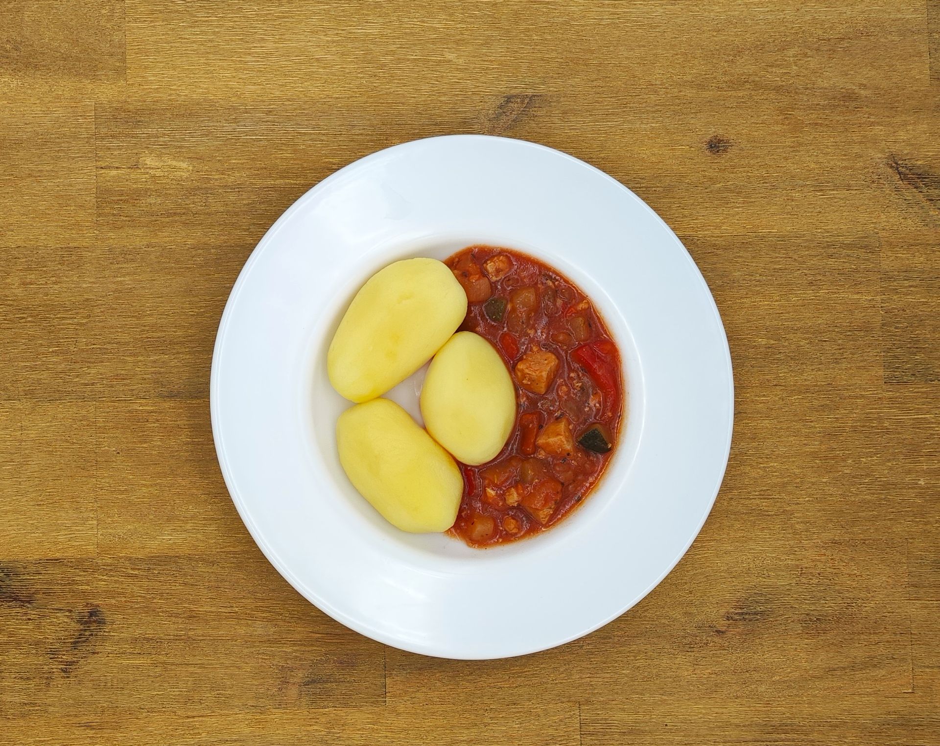 Weißer Teller mit drei gekochten Kartoffeln und einem Eintopf mit roter Soße auf einem Holztisch.