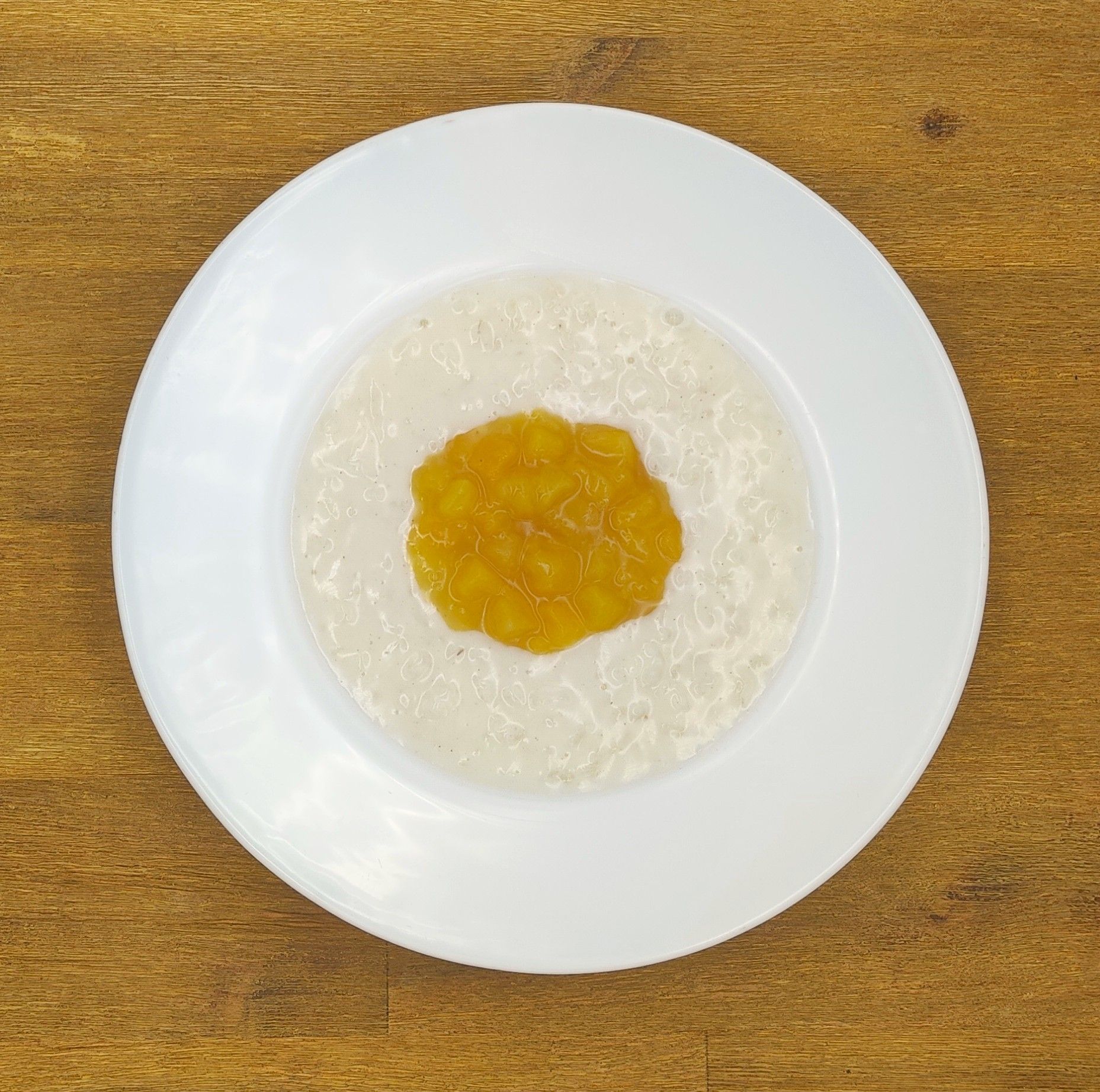 Weißer Teller mit cremefarbenem Haferbrei, garniert mit einem Klecks gelber Fruchtkompott, auf einer Holzoberfläche.