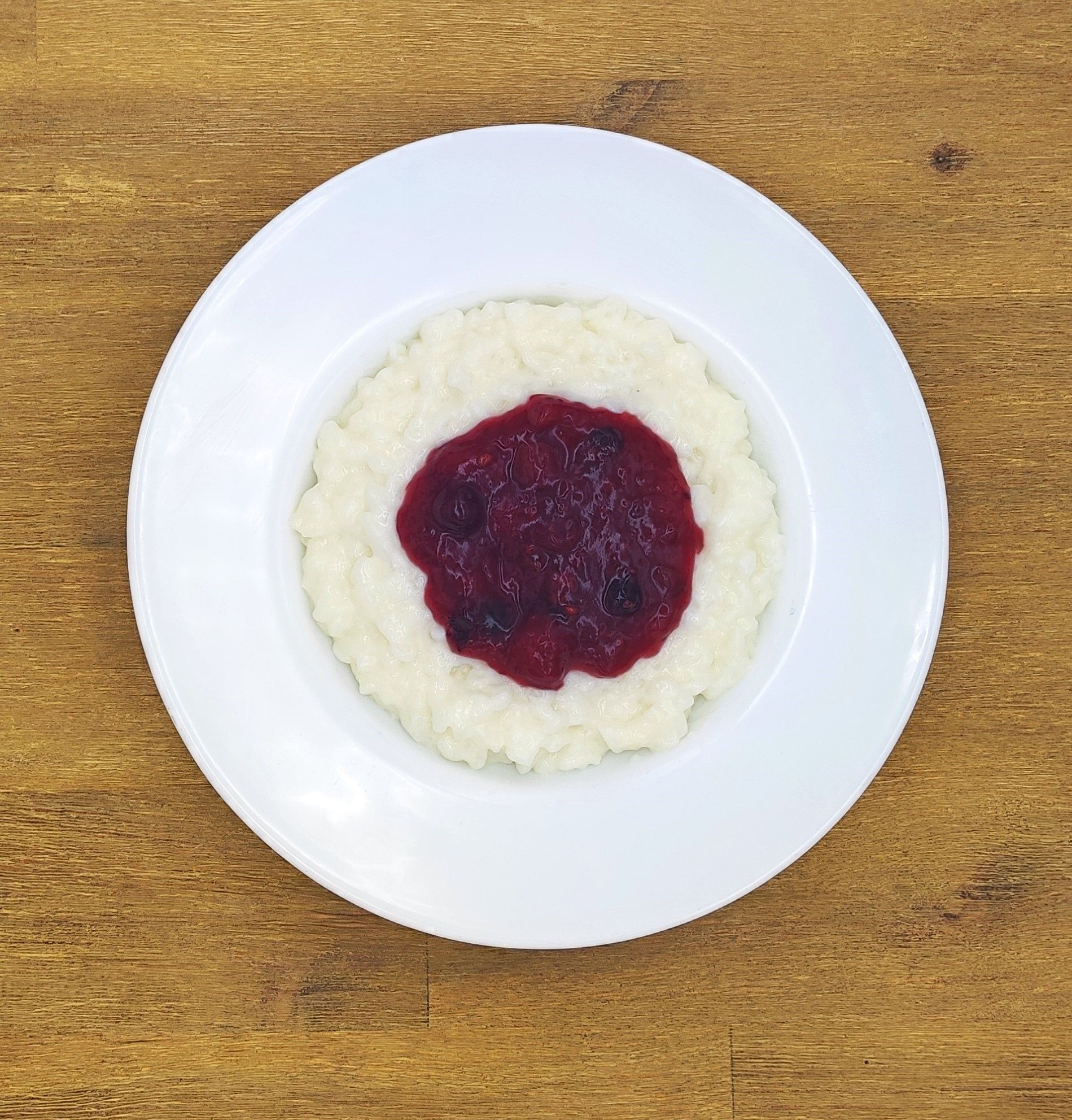 Weißer Milchreis mit einem Klecks roter Beerensauce in der Mitte, auf einem weißen Teller auf einer Holzoberfläche.
