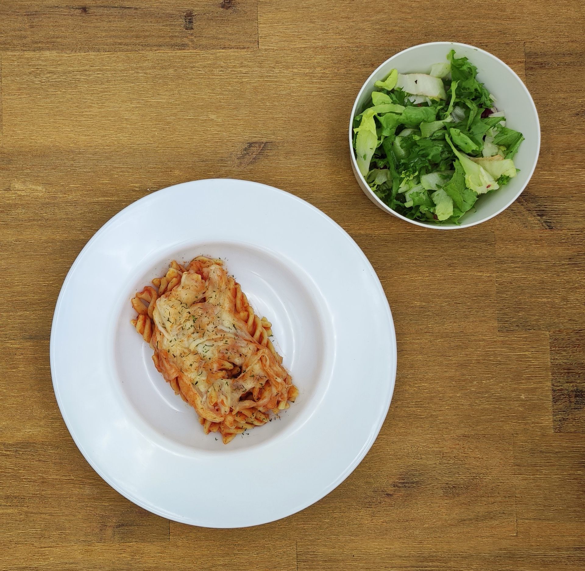 Nudelauflauf auf einem weißen Teller mit einem Beilagensalat in einer weißen Schüssel auf einem Holztisch.