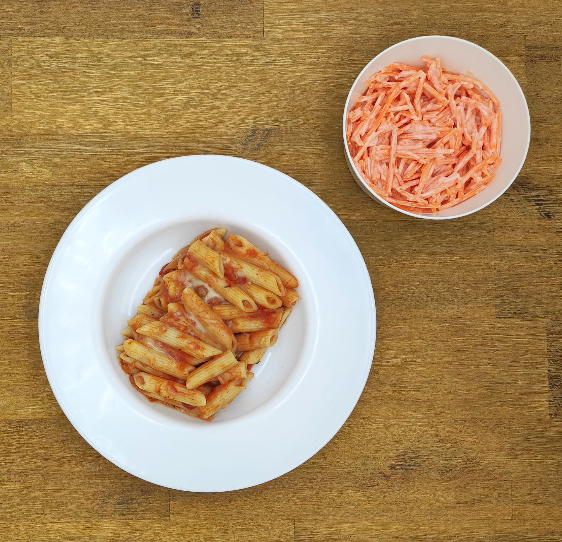 Teller Penne-Nudeln mit Tomatensoße, Schüssel Karottensalat auf einem Holztisch.