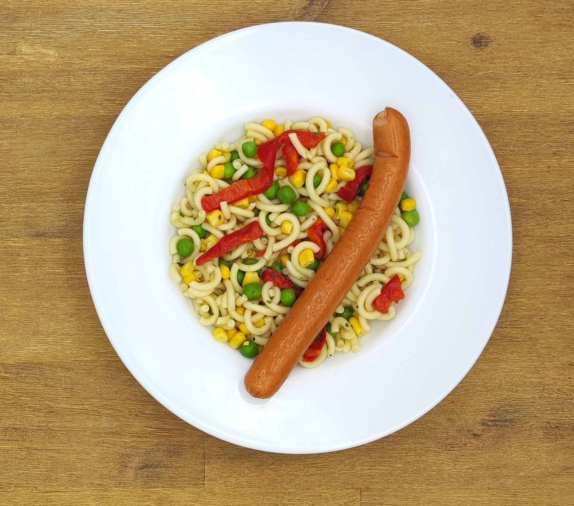 Teller mit Nudeln, Erbsen, Mais, roter Paprika und einer Wurst.