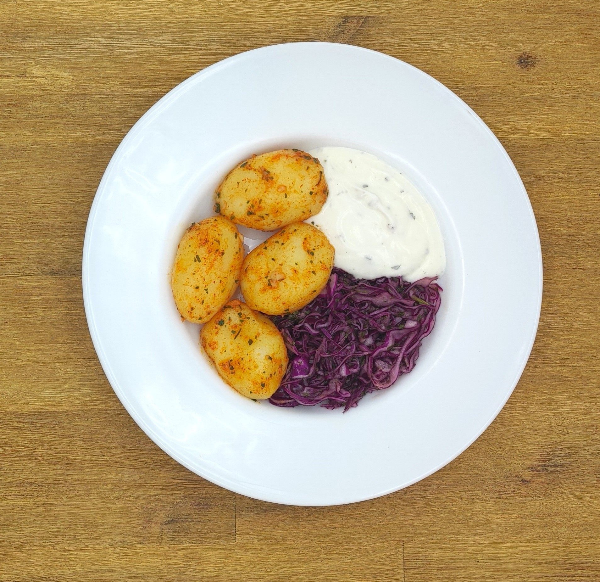 Ein Teller mit Bratkartoffeln, Rotkohl und weißer Soße auf einem Holztisch.