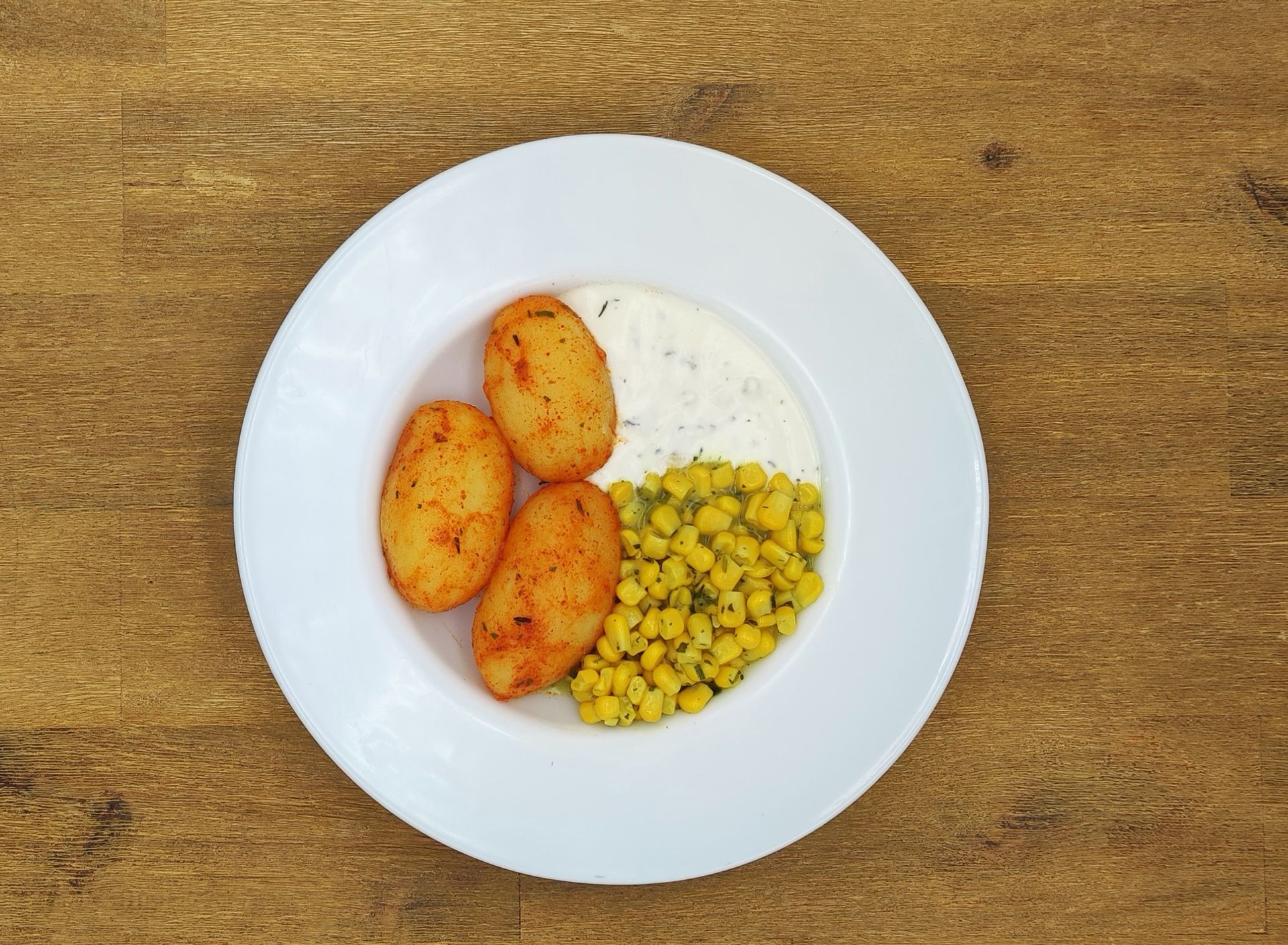 Weißer Teller mit drei gewürzten Kartoffeln, Mais und weißer Soße auf einer Holzoberfläche.