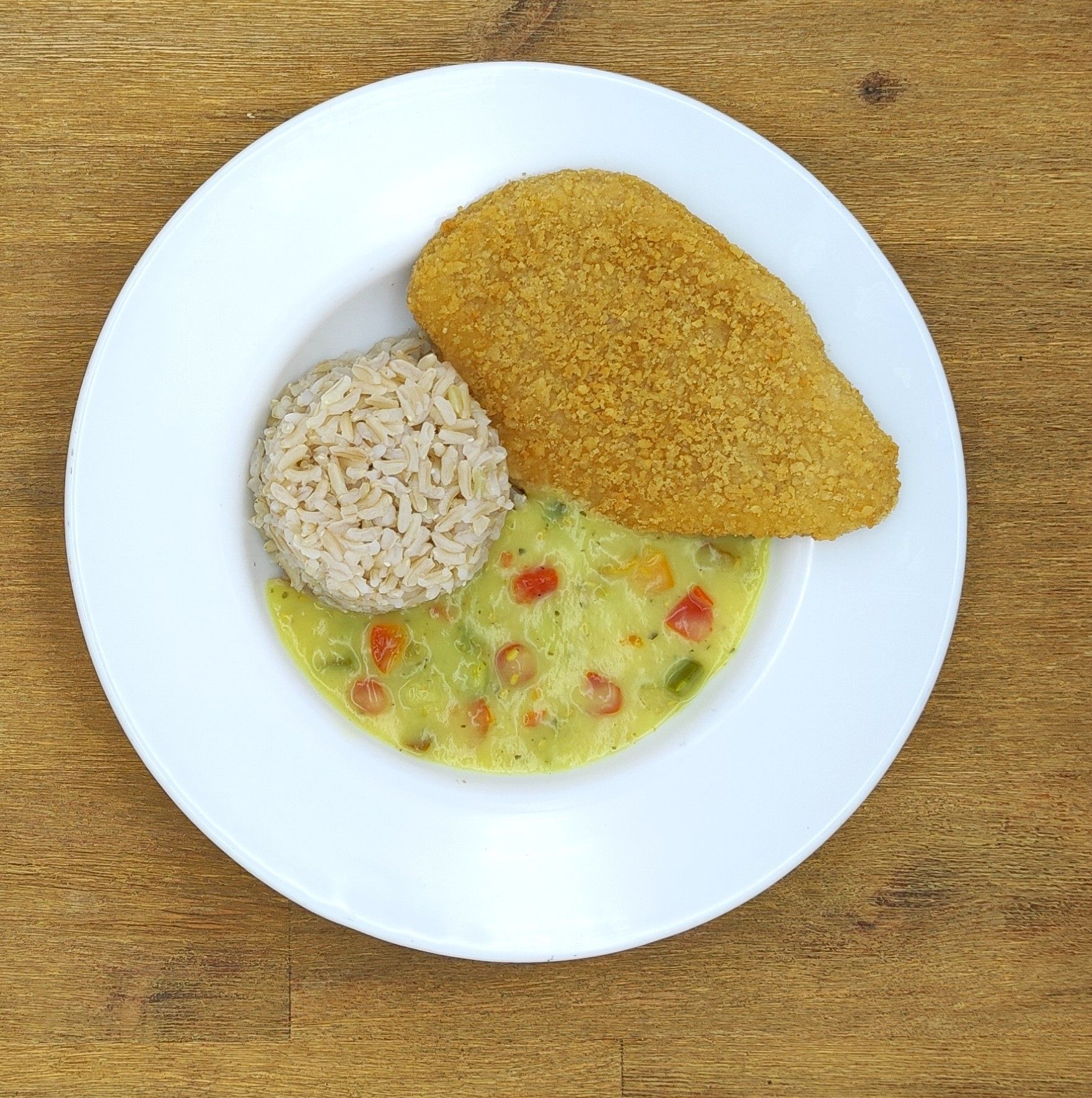 Paniertes Hähnchenbrustfilet, Reis und cremige Gemüsesauce auf einem weißen Teller, Holztisch.