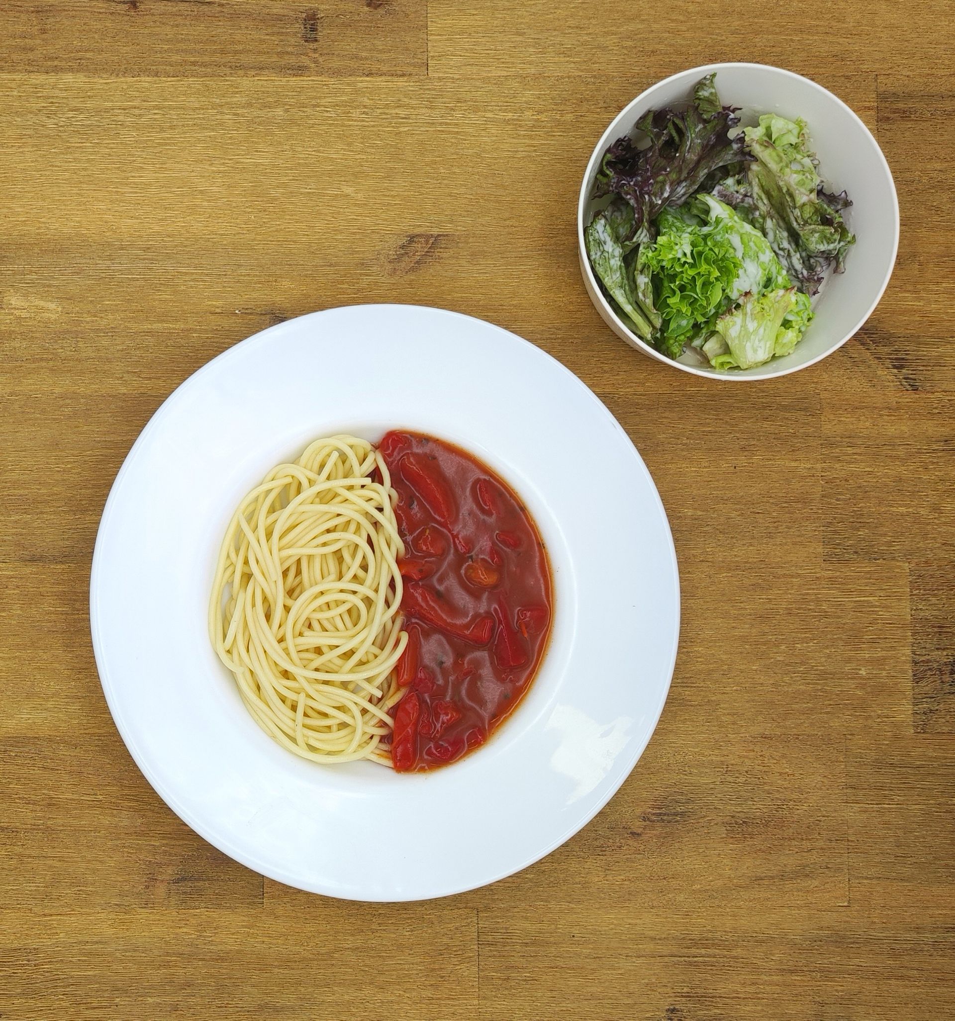 Spaghetti mit Tomatensoße und einem Beilagensalat in einer weißen Schüssel auf einer Holzoberfläche.
