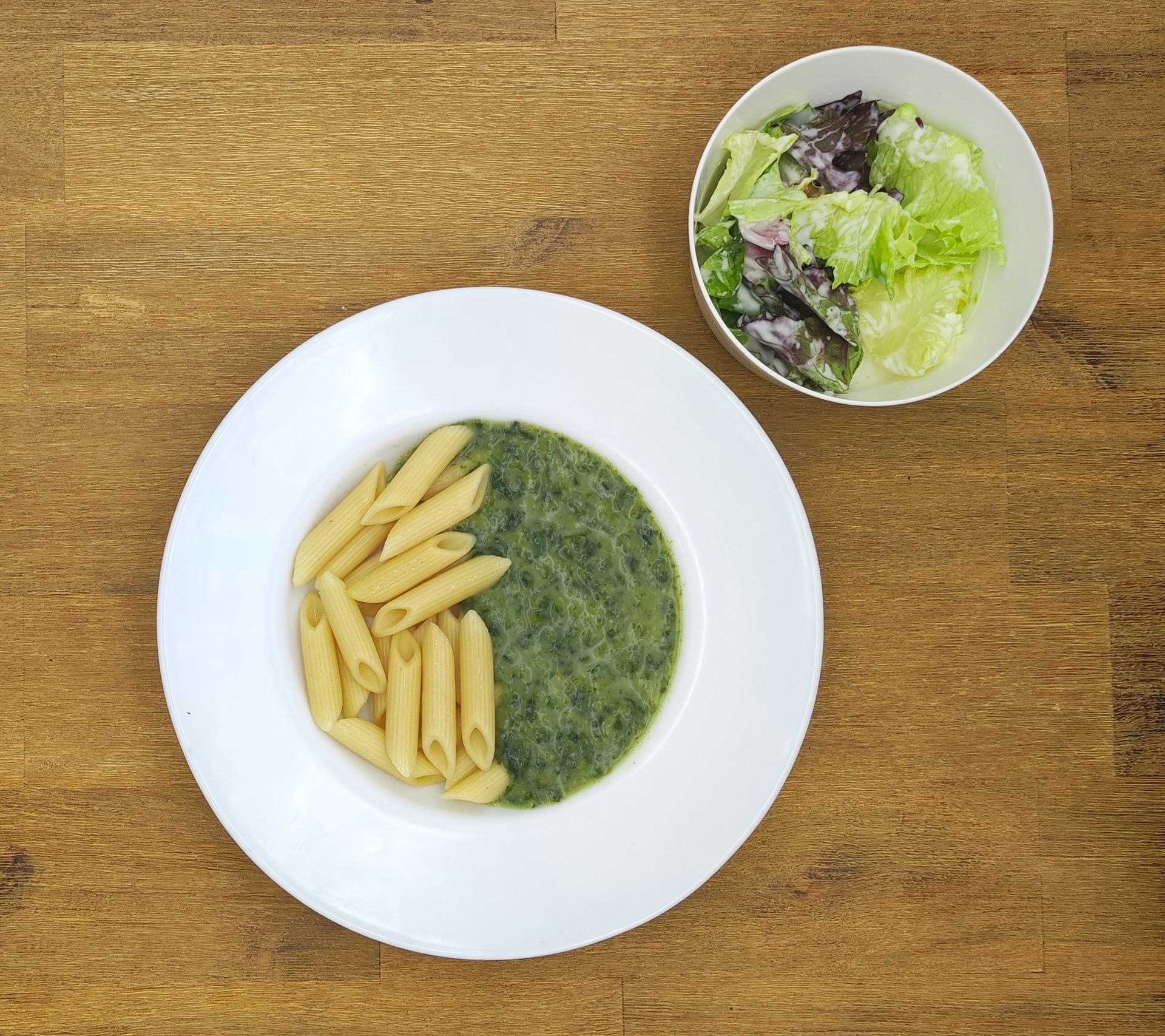 Pasta mit grüner Soße und einem Beilagensalat auf einem Holztisch.