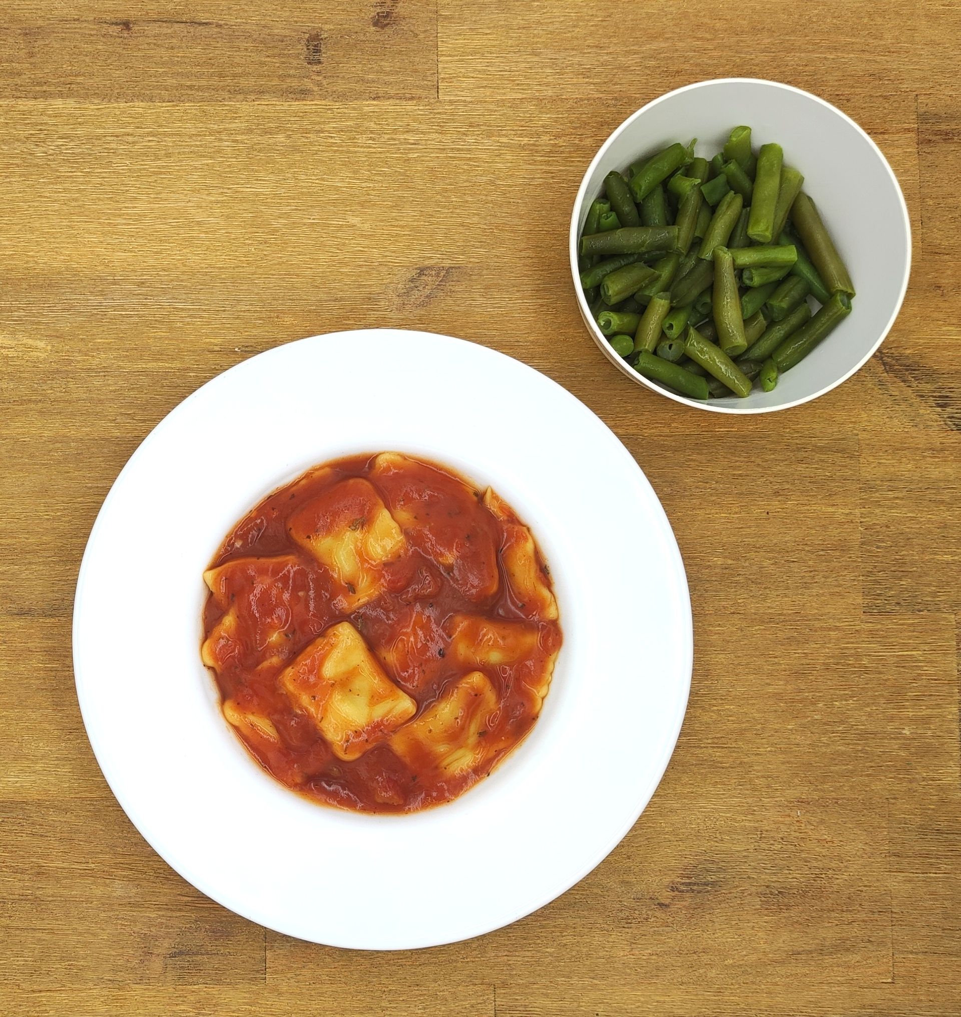 Ravioli in Tomatensoße auf einem weißen Teller, grüne Bohnen in einer weißen Schüssel, auf einer Holzoberfläche.