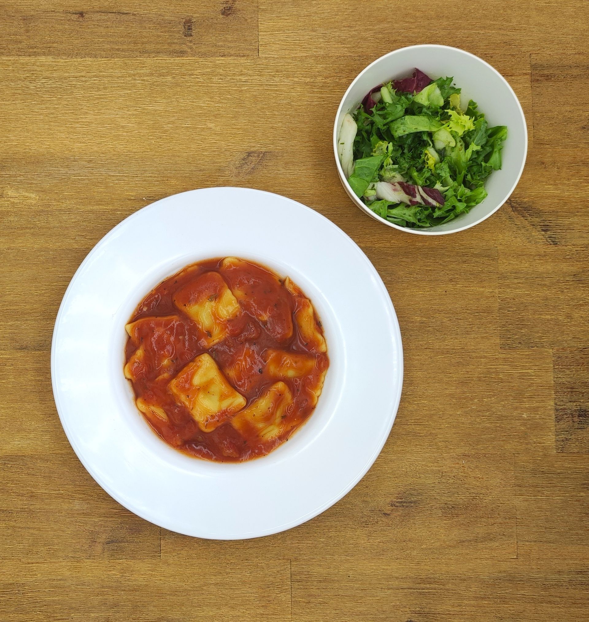 Pastagericht in Tomatensoße, dazu ein Salat in einer weißen Schüssel, auf einem Holztisch.