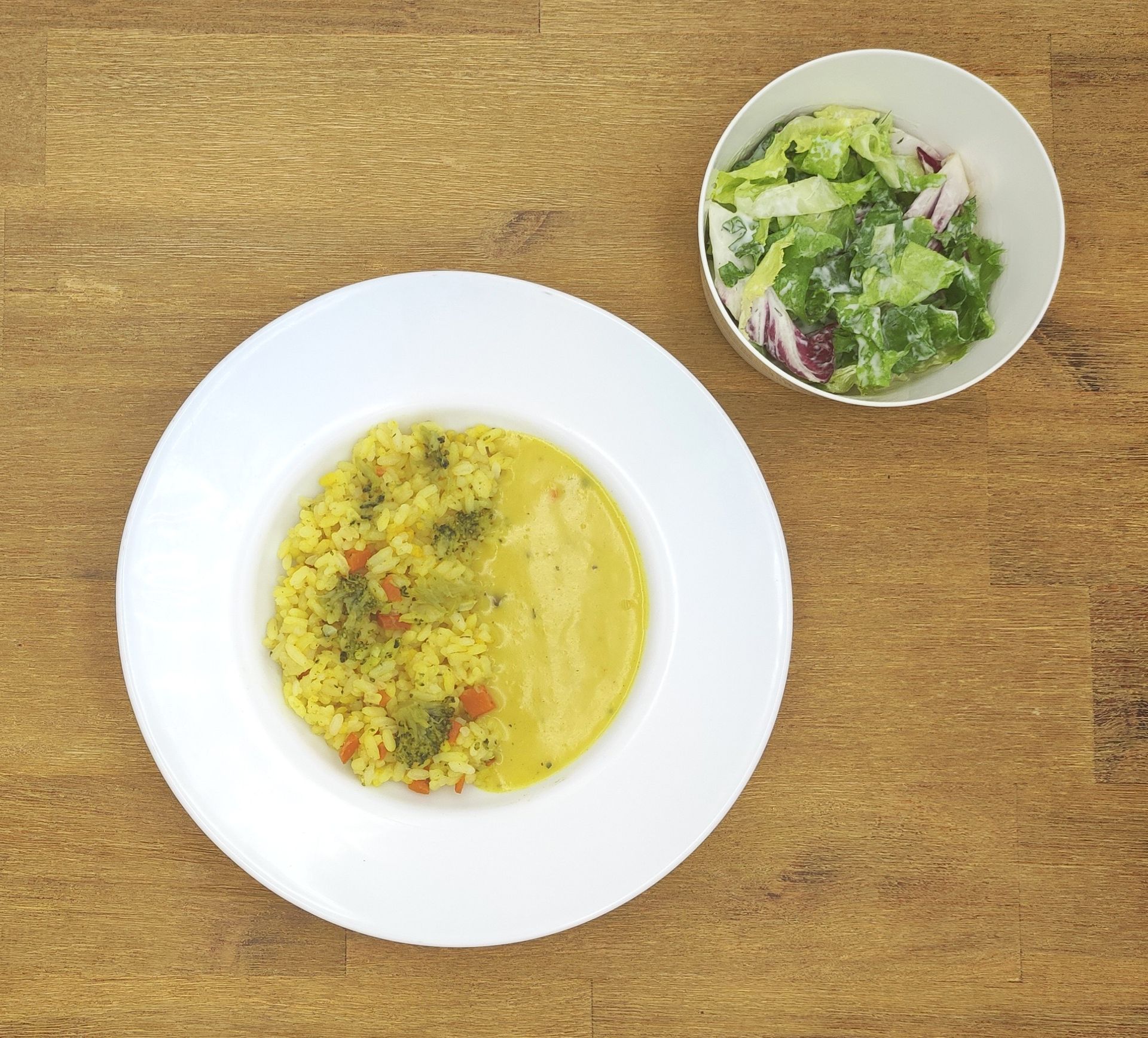 Ein Teller mit gelbem Reis und Soße, dazu ein Beilagensalat, auf einem Holztisch.