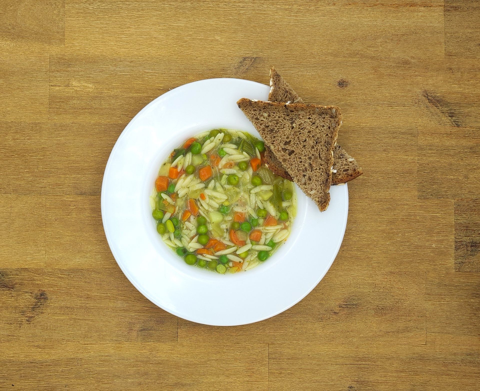 Eine weiße Schüssel mit Orzo-Gemüsesuppe und zwei Scheiben dunklem Brot auf einem Holztisch.