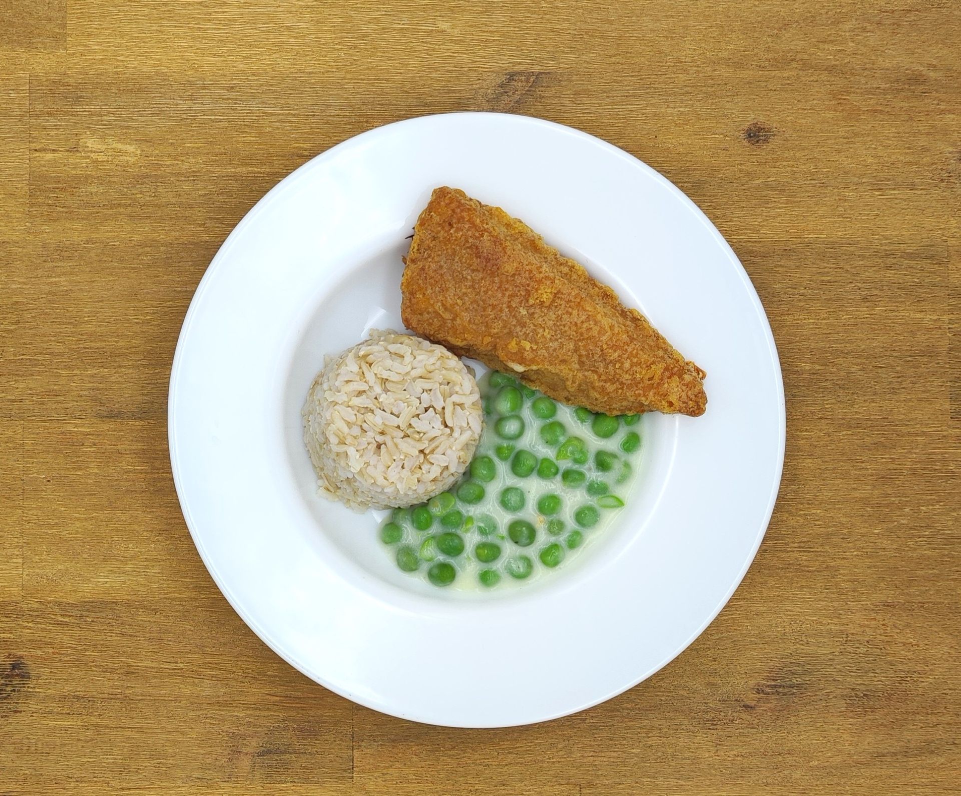 Ein Teller mit paniertem Fisch, Reis und Erbsen auf einer Holzoberfläche.