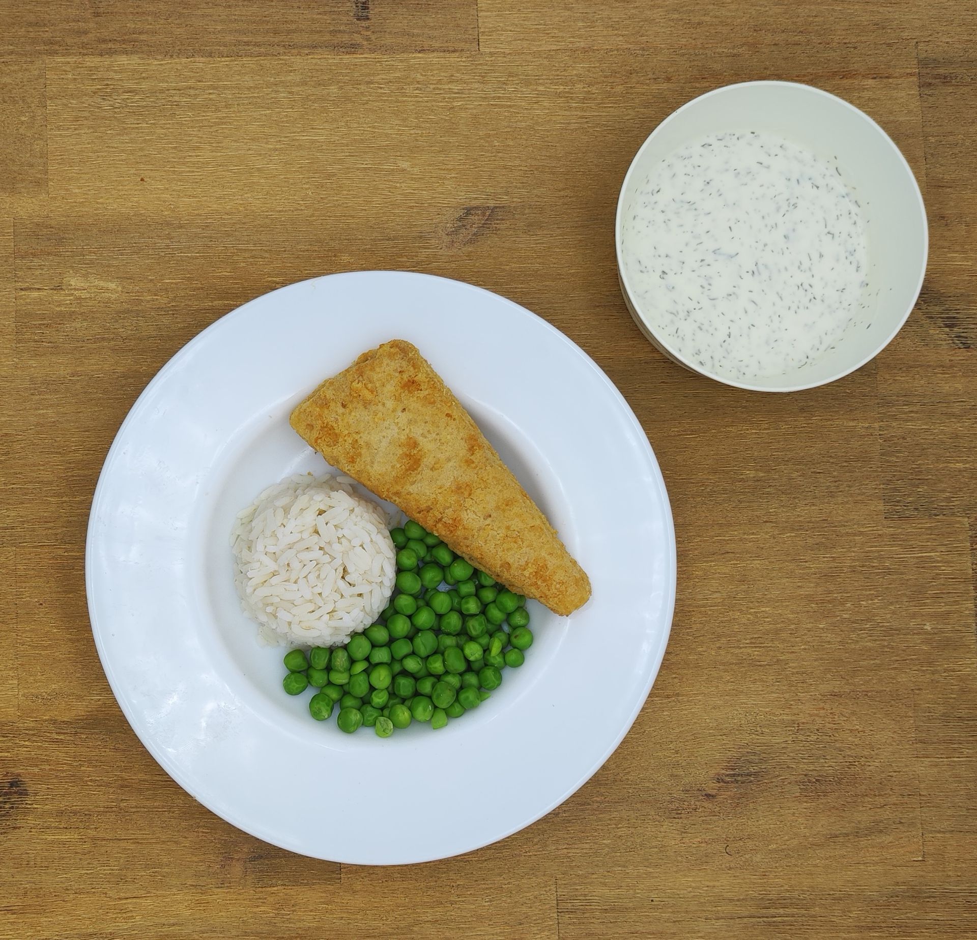 Teller mit Fisch, Erbsen und Reis; kleine Schüssel mit Reis auf einer Holzoberfläche.