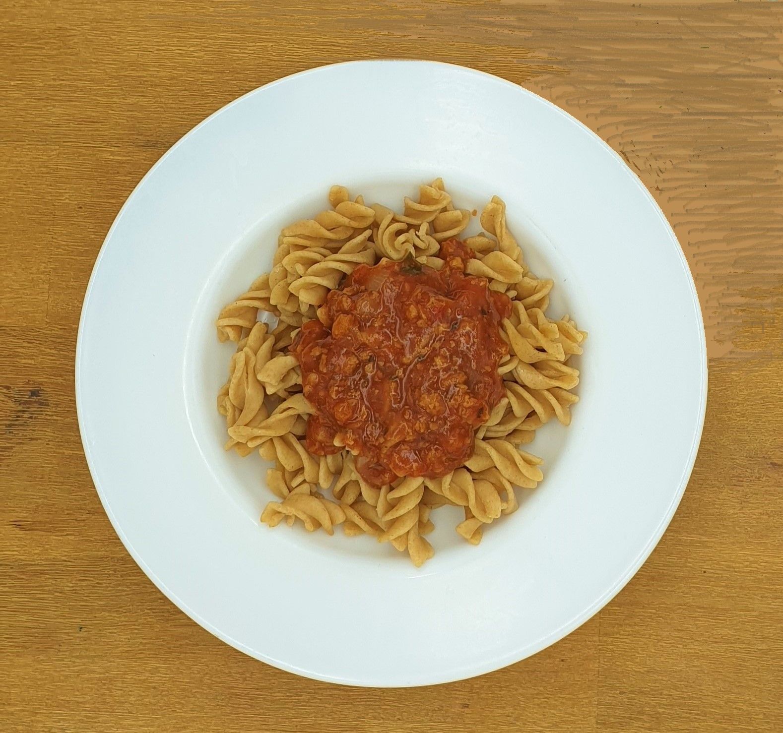 Ein Teller mit Vollkornnudeln in roter Soße, zentriert auf einem hellen Holztisch.