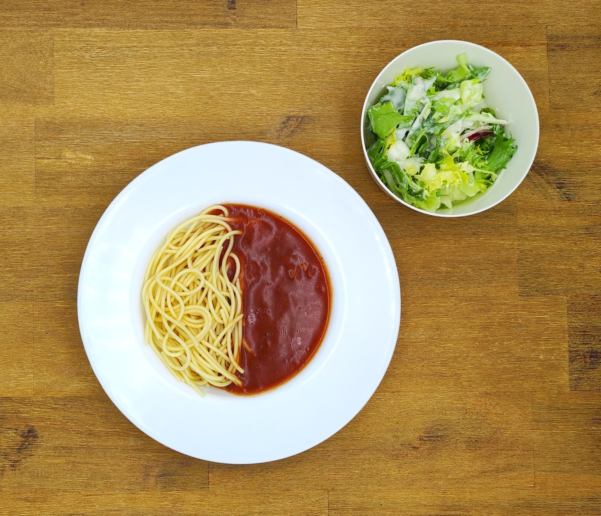Spaghetti mit Tomatensoße auf einem weißen Teller, Salat in einer Schüssel auf einem Holztisch.