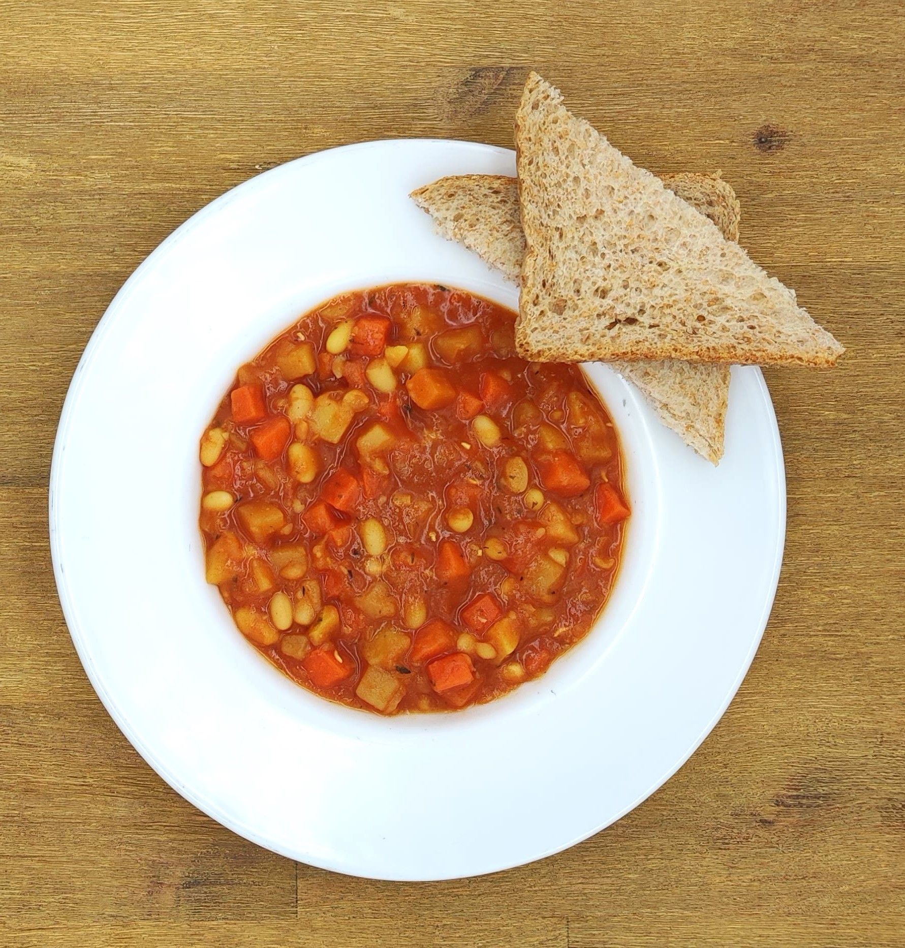 Weiße Schüssel mit Gemüsesuppe und Brot auf einem Holzbrett.