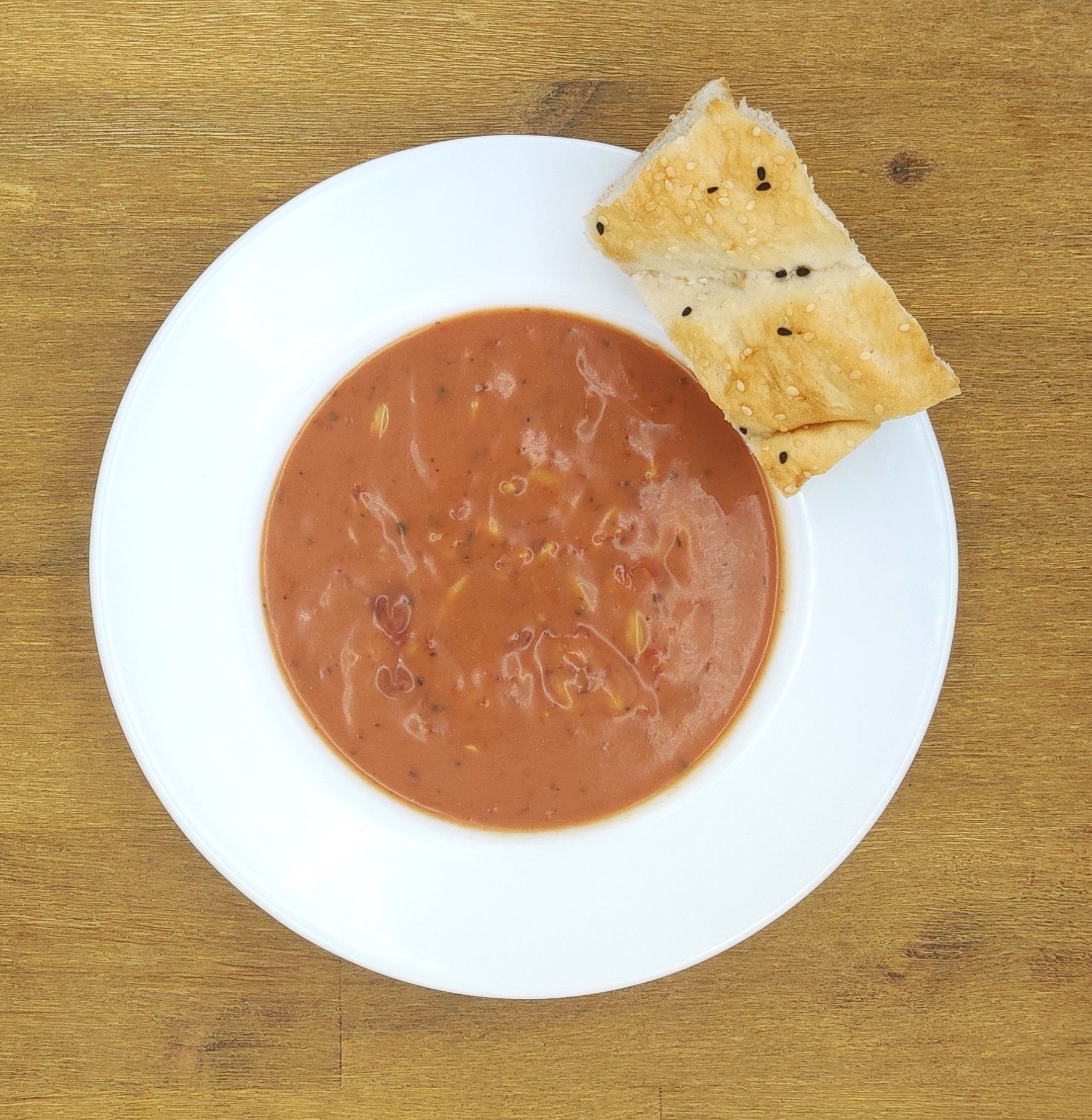 Weiße Schüssel mit rötlicher Suppe und einem Stück Fladenbrot auf einer Holzoberfläche.