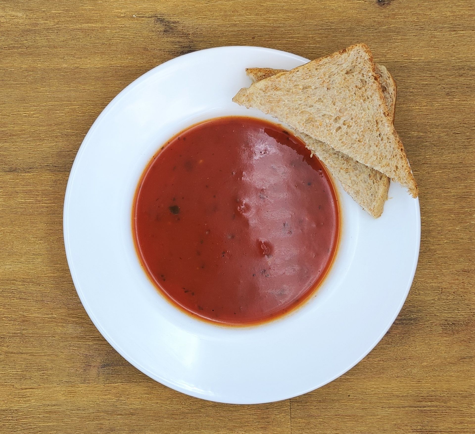 Eine Schüssel Tomatensuppe mit zwei Scheiben Brot auf einem weißen Teller, Holztisch.