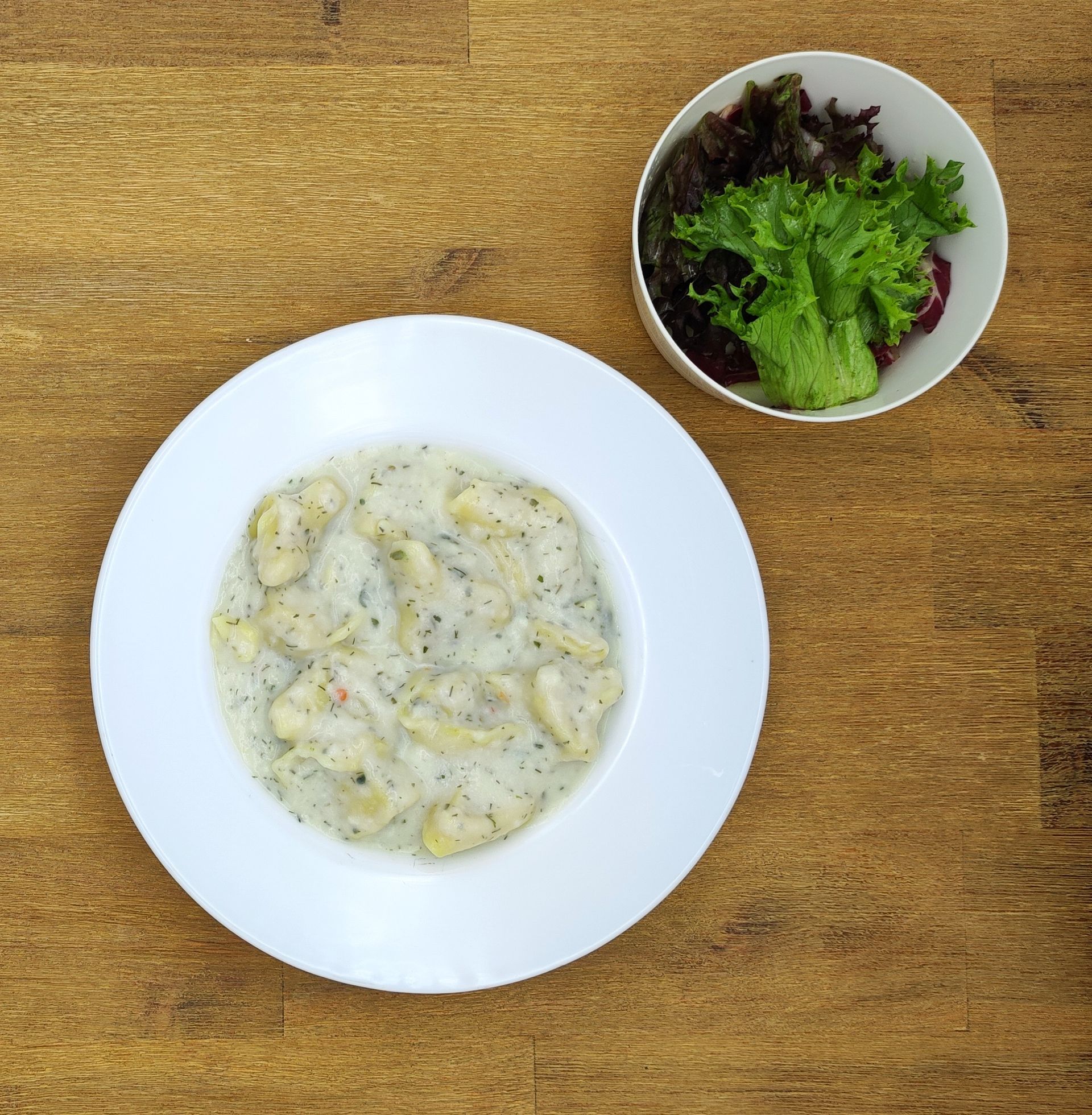 Ein weißer Teller mit Gnocchi in cremiger Soße neben einer Schüssel Salat auf einem Holztisch.
