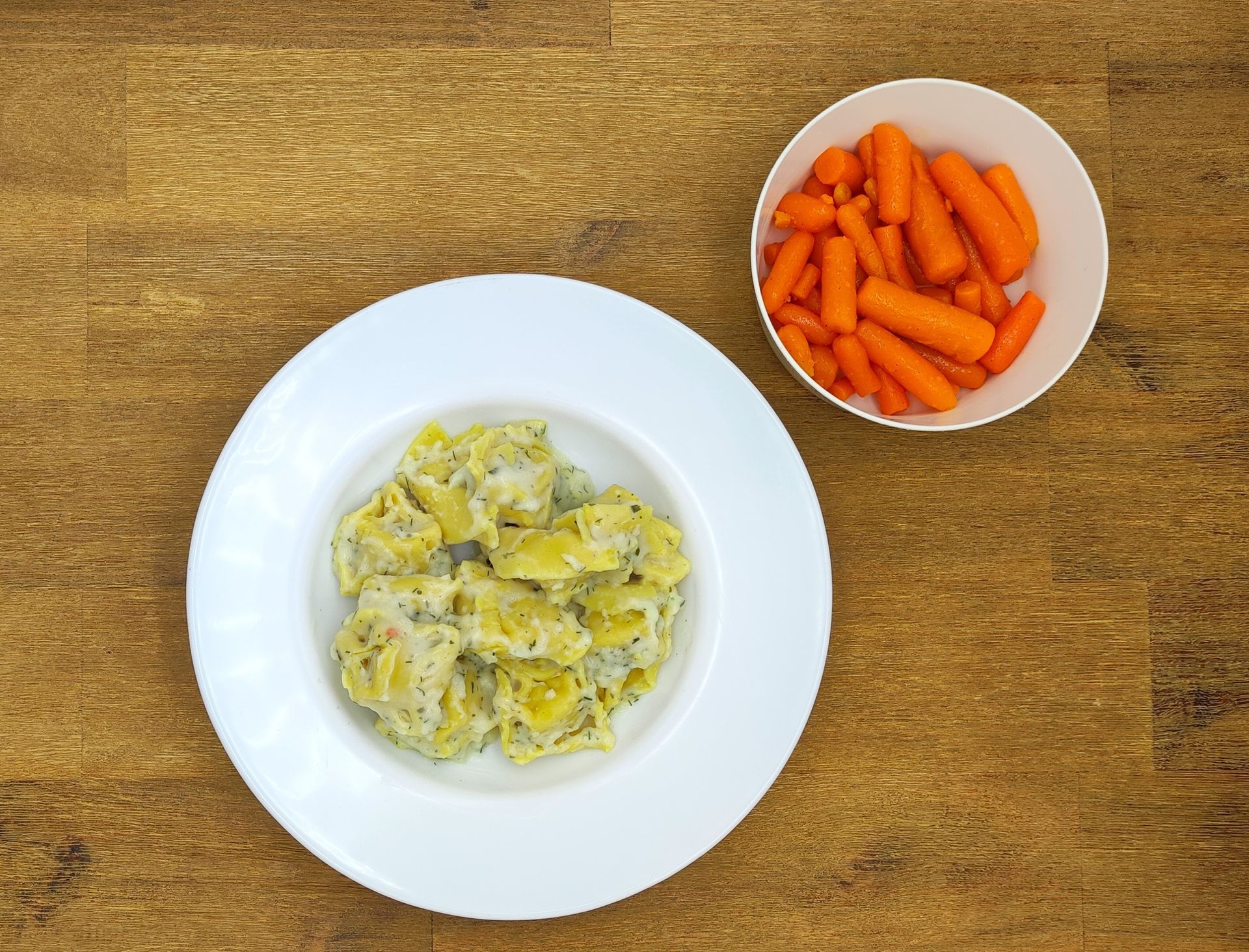 Kartoffelsalat auf einem weißen Teller und eine Schüssel mit Babykarotten auf einem Holztisch.
