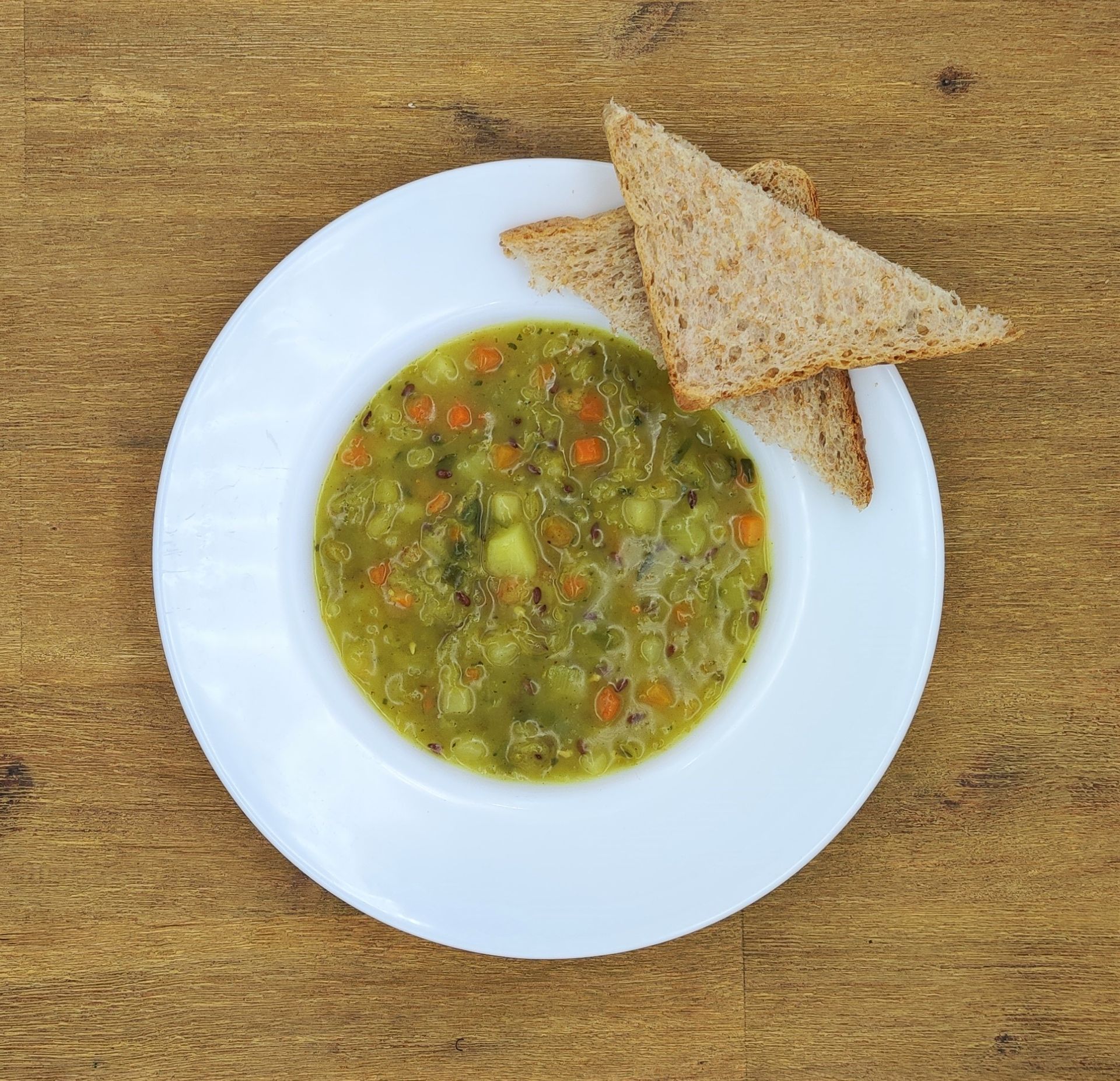 Eine Schüssel grüne Suppe mit Gemüse und zwei Scheiben Brot auf einem weißen Teller, der auf einer Holzoberfläche steht.