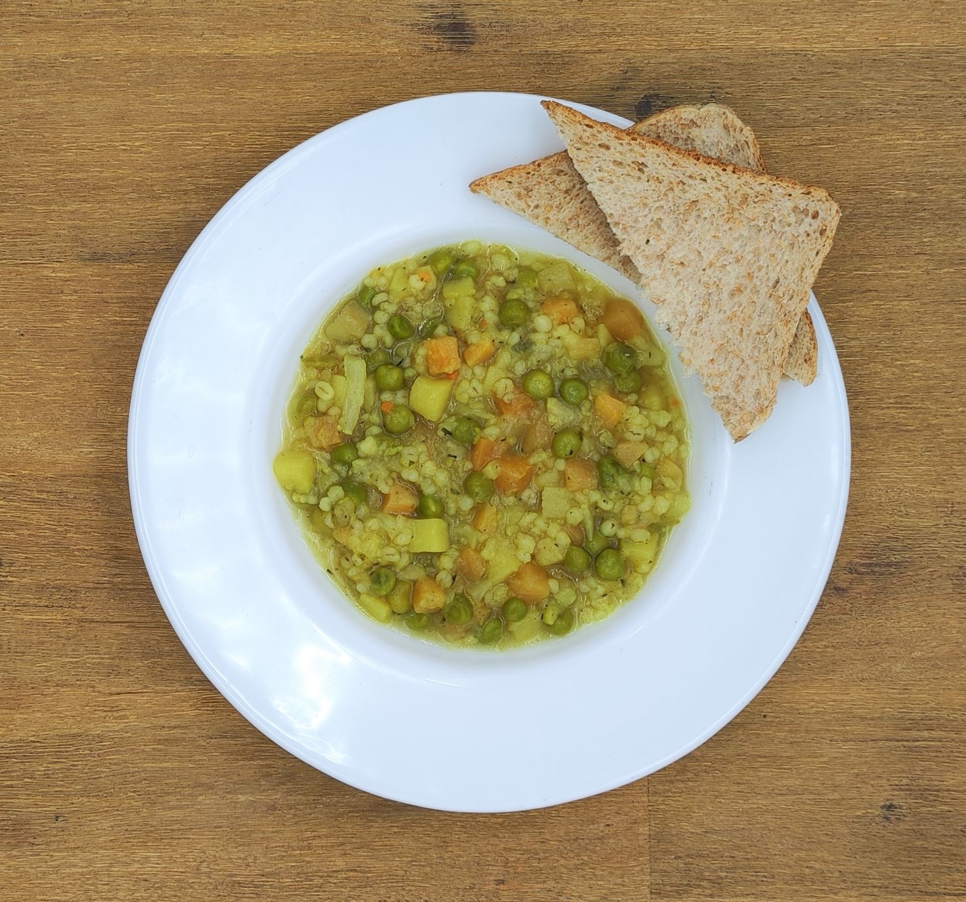 Gemüsesuppe, serviert auf einem weißen Teller mit zwei Scheiben geröstetem Vollkornbrot auf einem Holztisch.