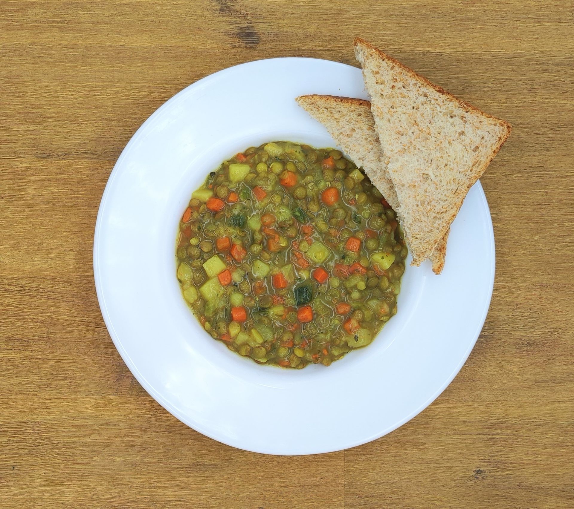 Weiße Schüssel mit Linsensuppe und Brot auf einer Holzoberfläche.