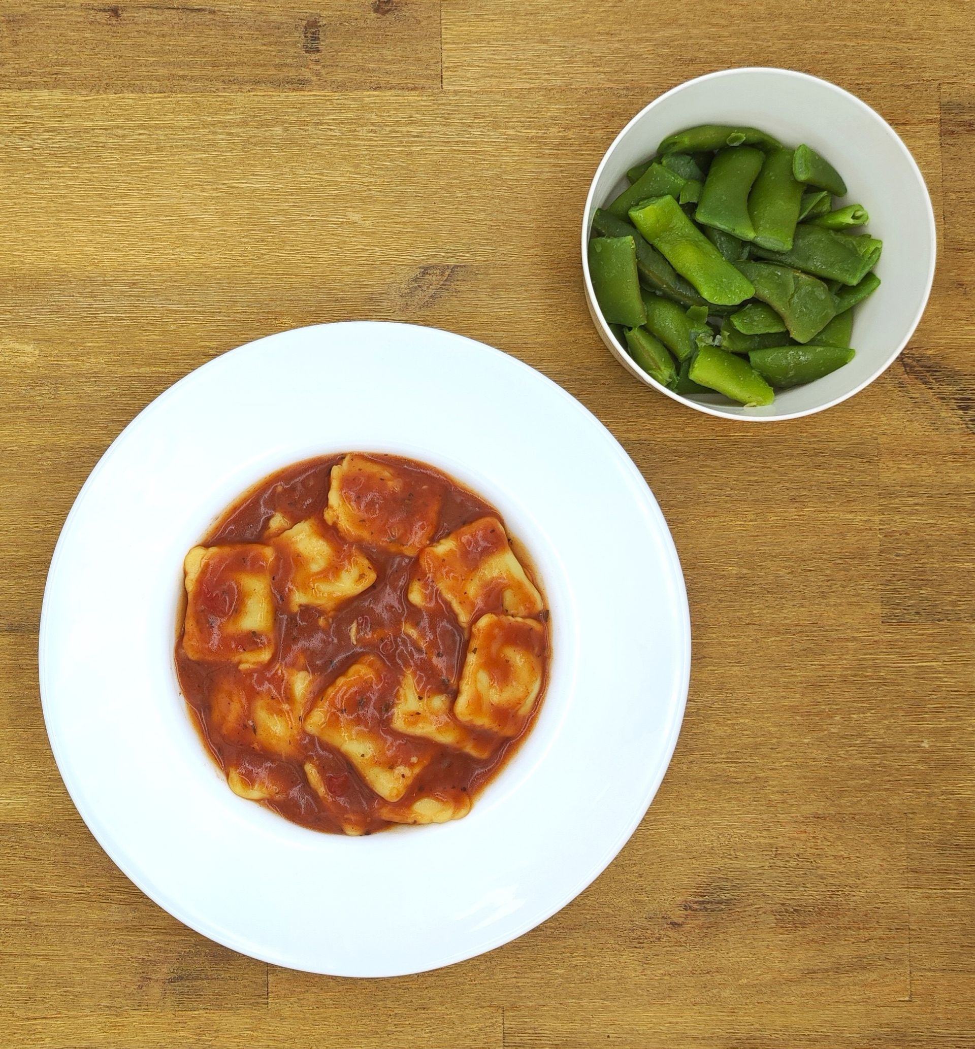 Ein Teller Ravioli in roter Soße neben einer Schüssel mit grünem Gemüse auf einer Holzoberfläche.