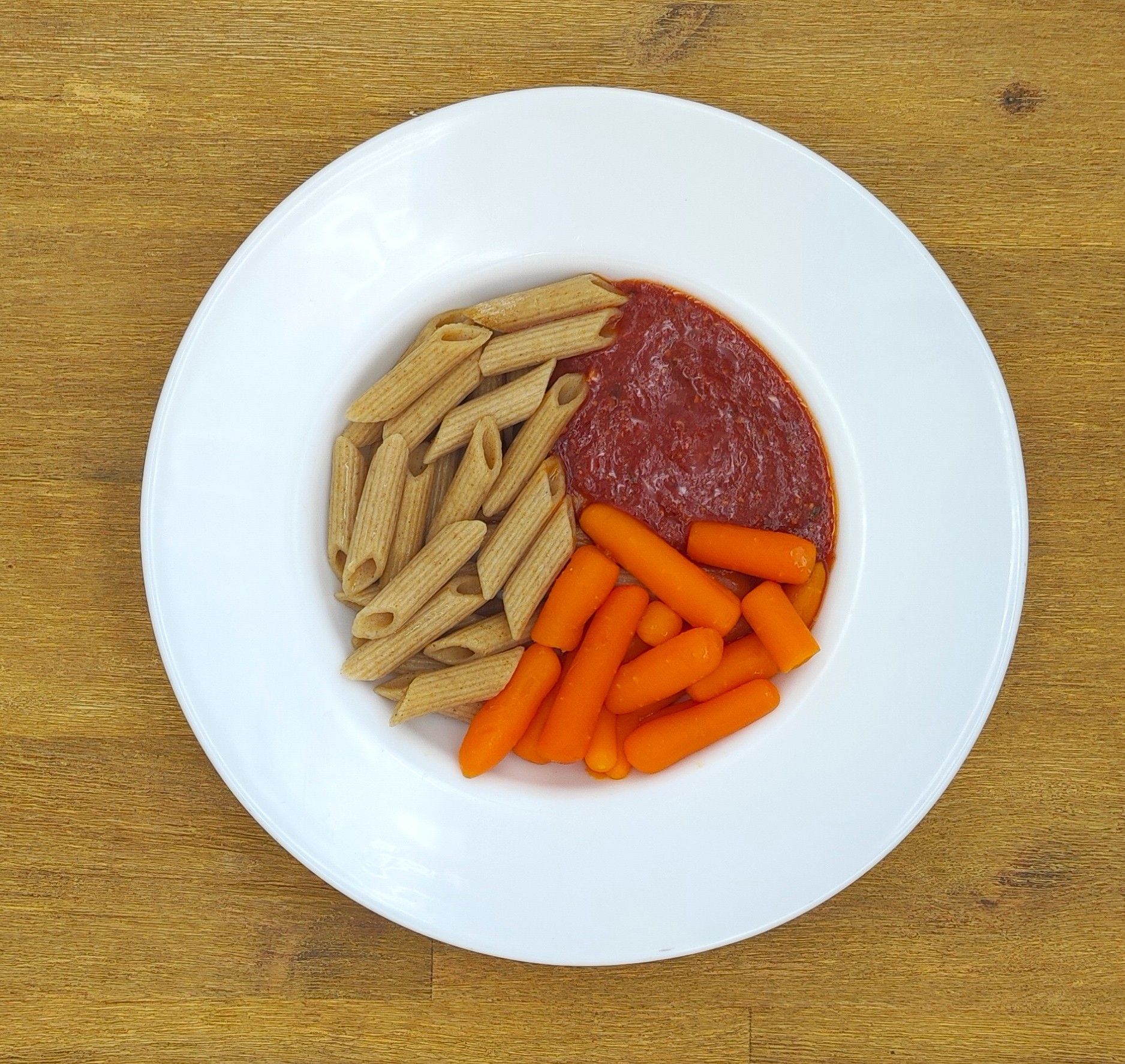 Ein Teller mit Nudeln, Tomatensoße und Babykarotten auf einem Holztisch.