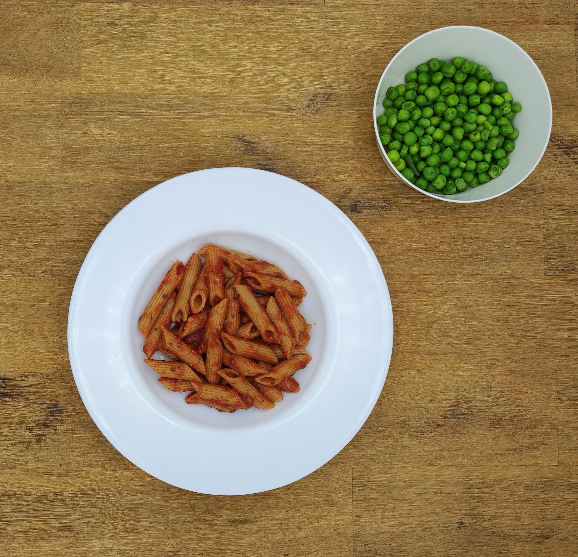 Pasta mit roter Soße auf einem weißen Teller, Erbsen in einer weißen Schüssel, auf einem Holztisch.