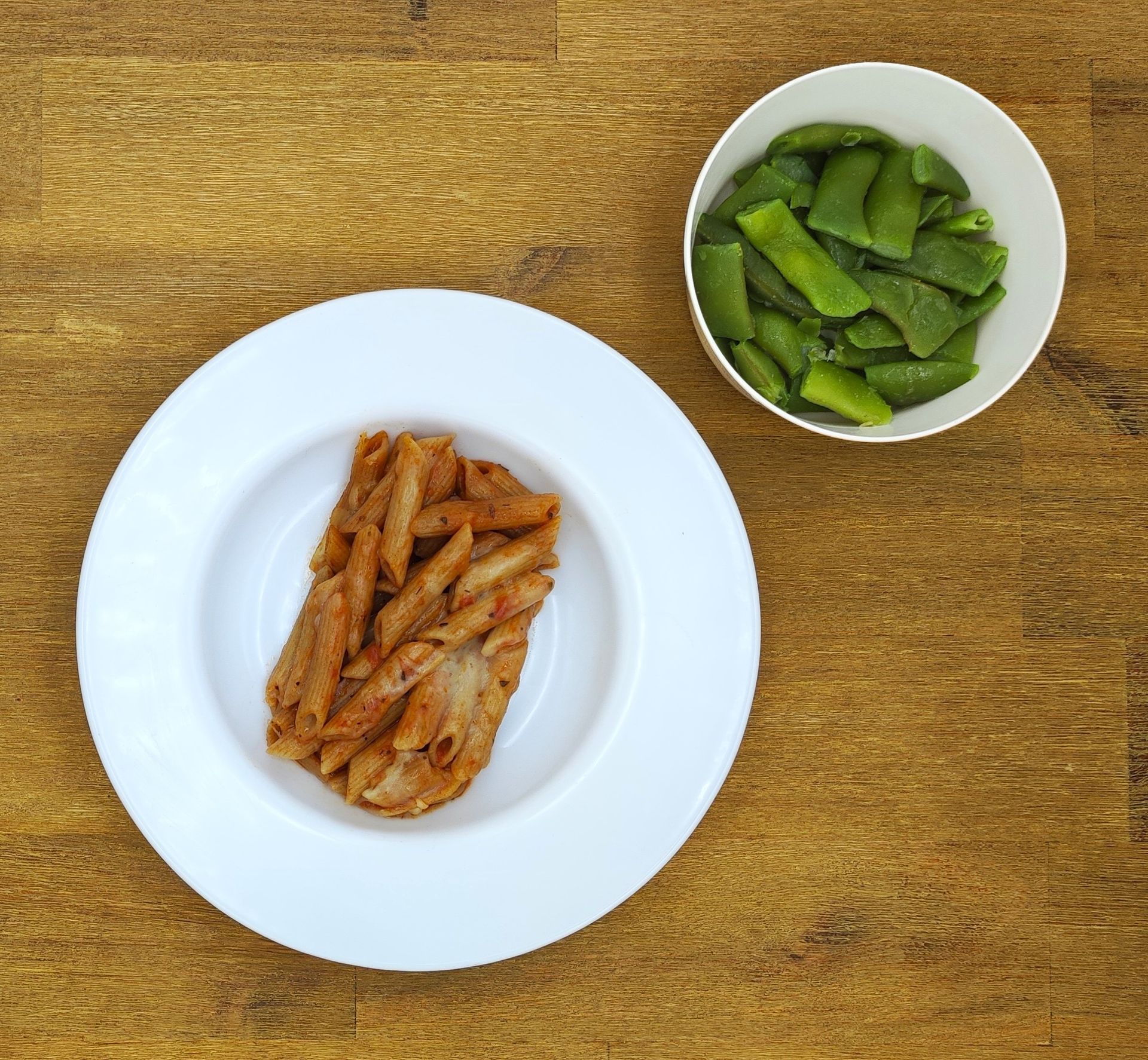 Pasta mit roter Soße auf einem weißen Teller, daneben eine Schüssel mit grünen Bohnen auf einer Holzplatte.