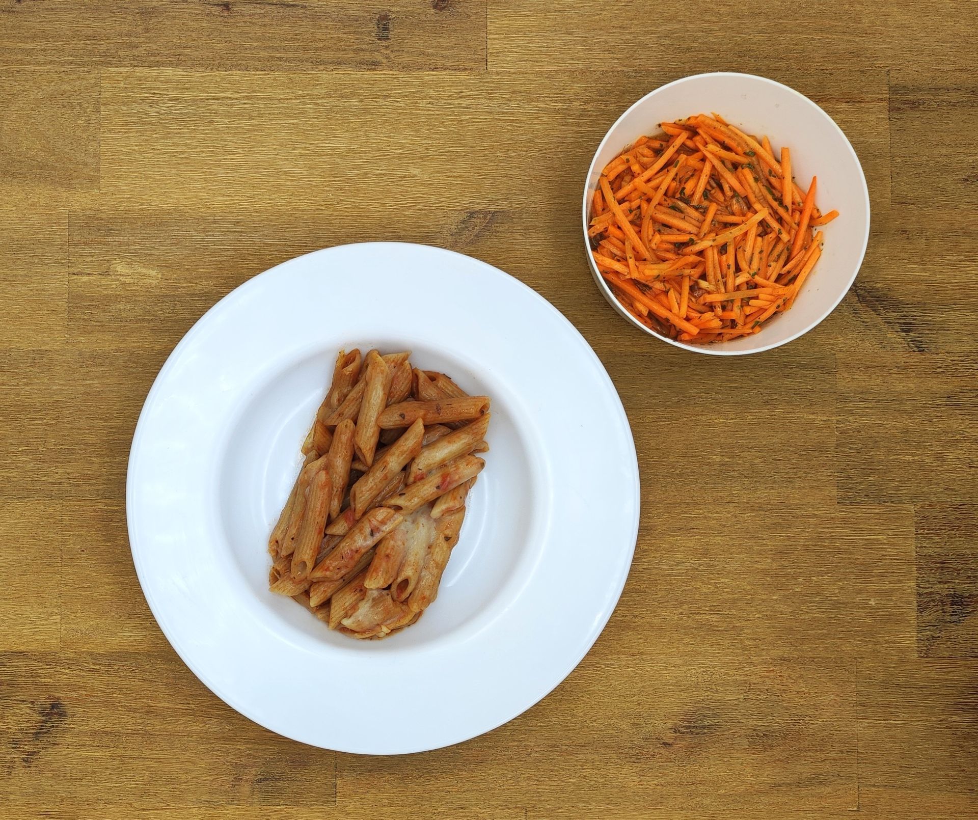 Pasta mit Soße auf einem weißen Teller; Karottensalat in einer weißen Schüssel, auf einer Holzoberfläche angerichtet.