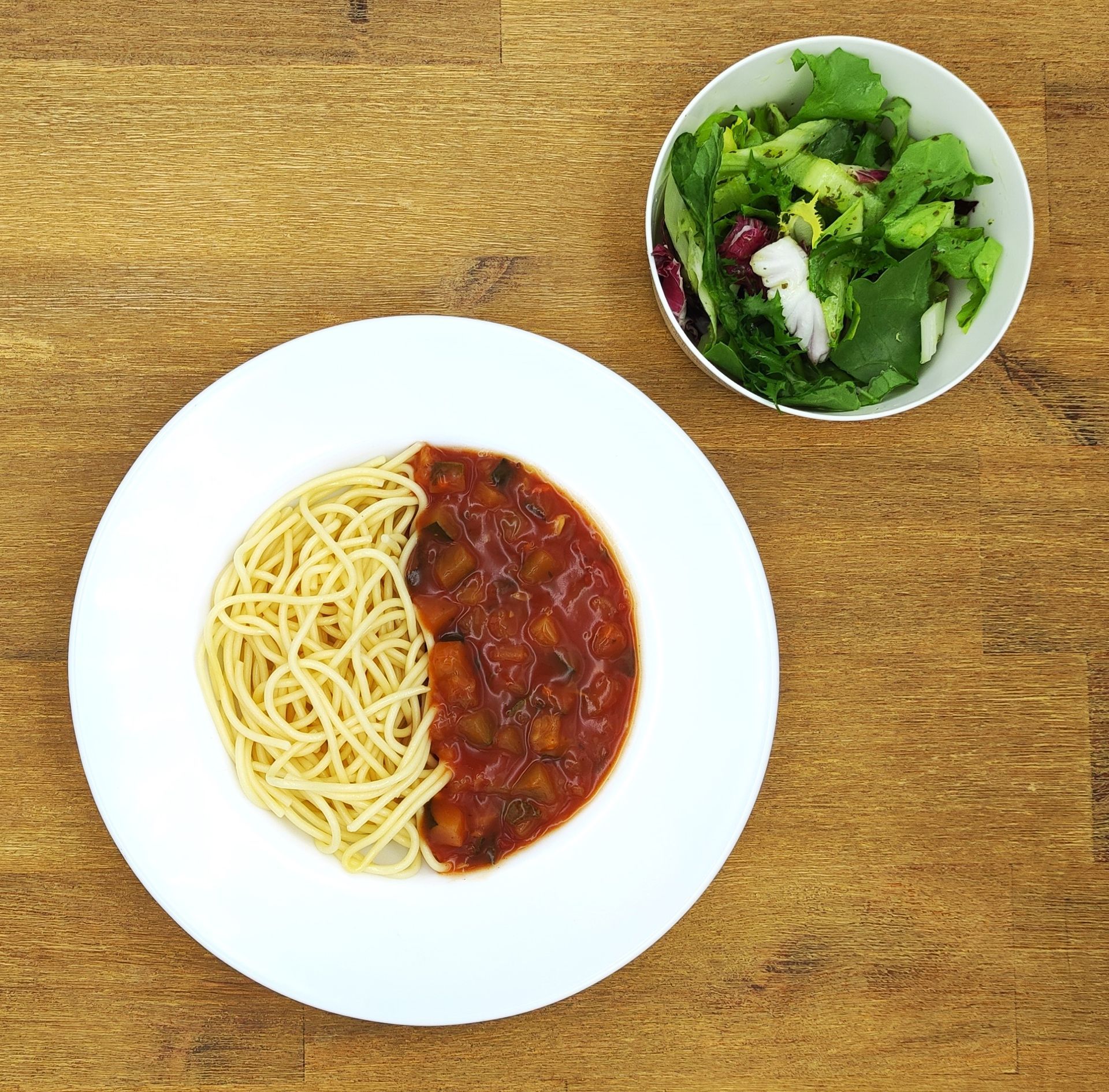 Spaghetti mit Tomatensoße auf einem weißen Teller neben einer kleinen Schüssel Salat auf einem Holztisch.