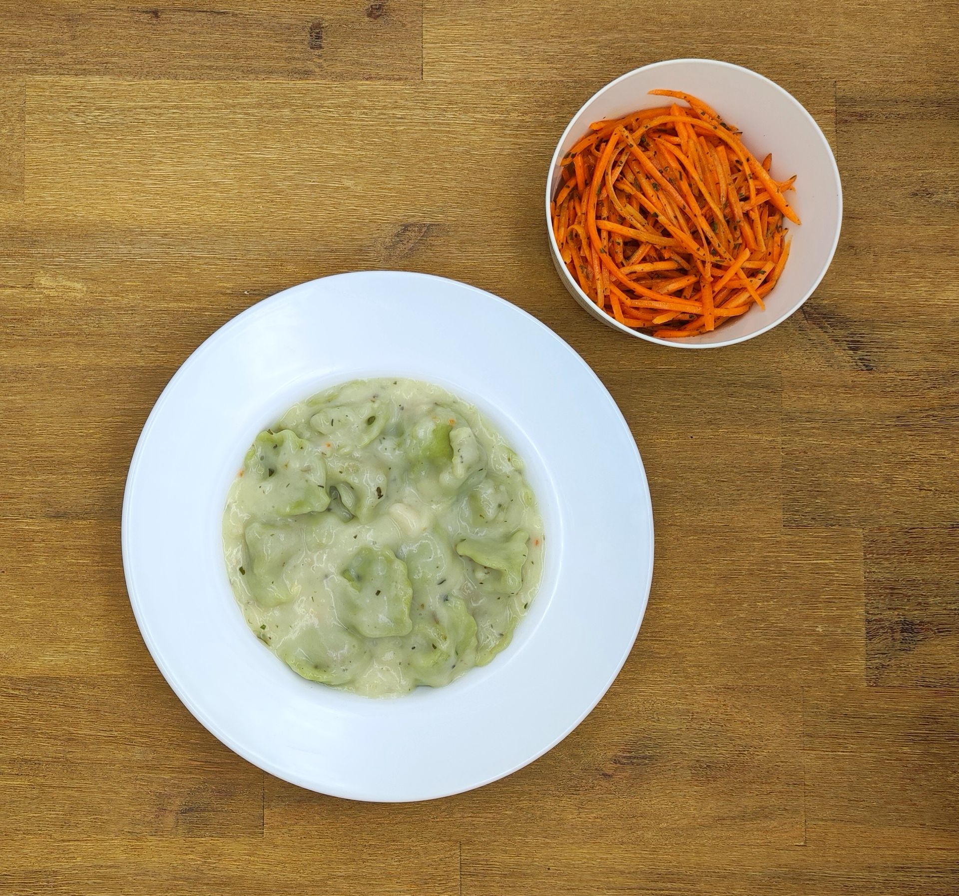 Ein Teller mit Ravioli und eine Schüssel mit geraspelten Karotten auf einem Holztisch.