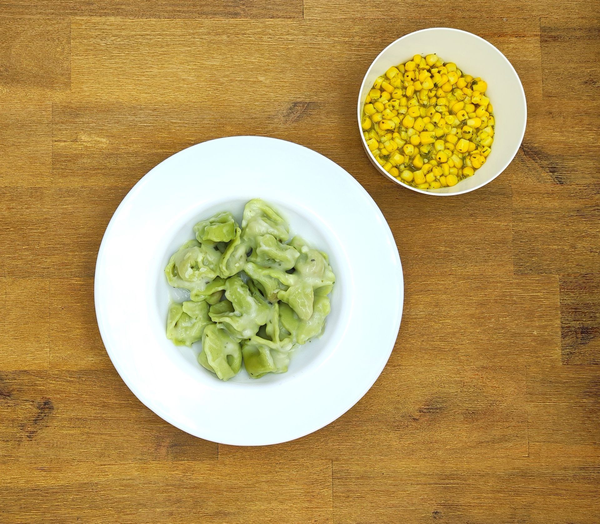 Pasta mit grüner Soße auf einem weißen Teller und eine Schüssel Mais auf einem Holztisch.