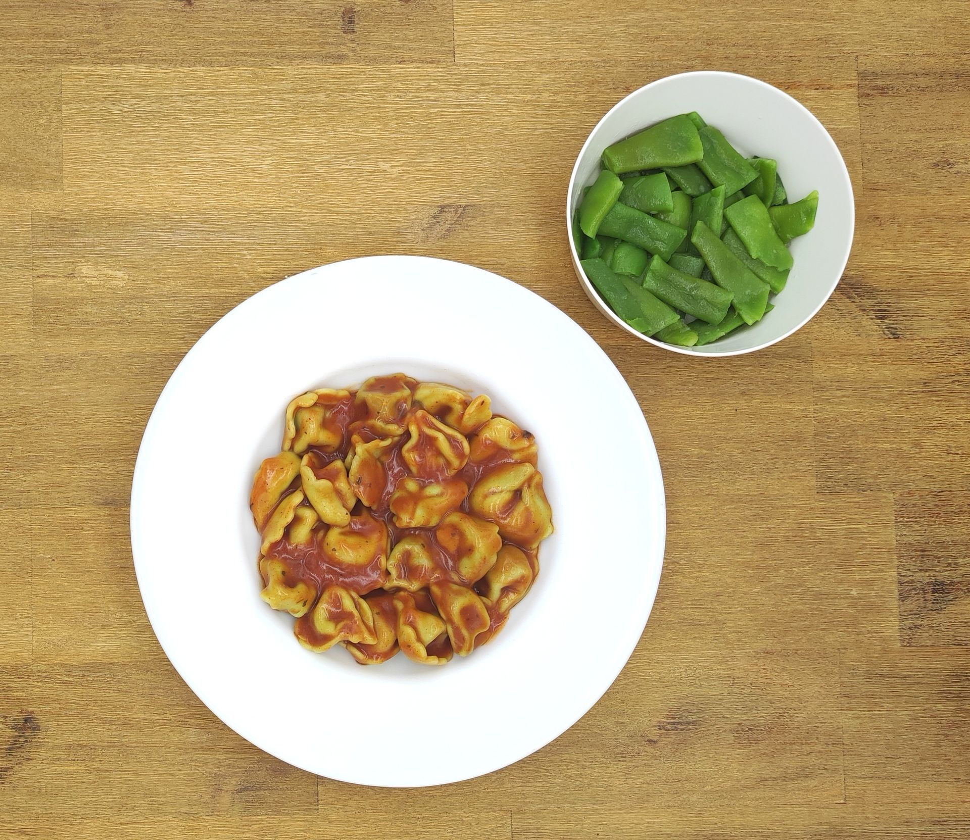 Tortellini mit roter Soße in einer weißen Schüssel, Zuckerschoten in einer separaten Schüssel, auf einer Holzoberfläche.