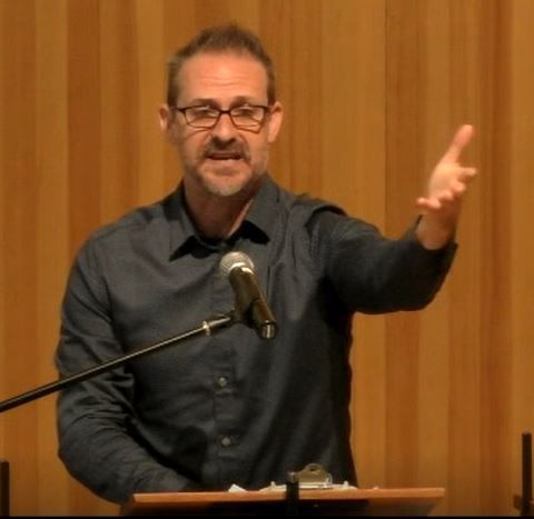 Chris Crass speaking at the pulpit with his left arm outstretched