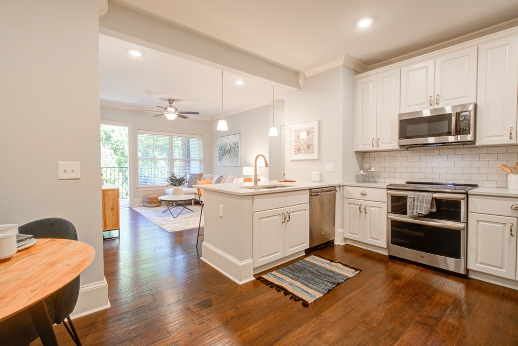 Open-concept kitchen with white cabinets, stainless steel appliances, and island. Dark wood floors lead to a living area.