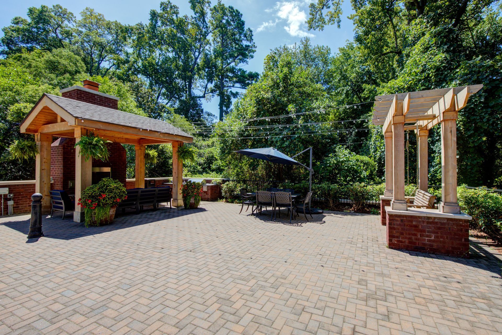 Brick patio with a pergola, covered seating area with fireplace, and table with umbrella. Green trees in background.